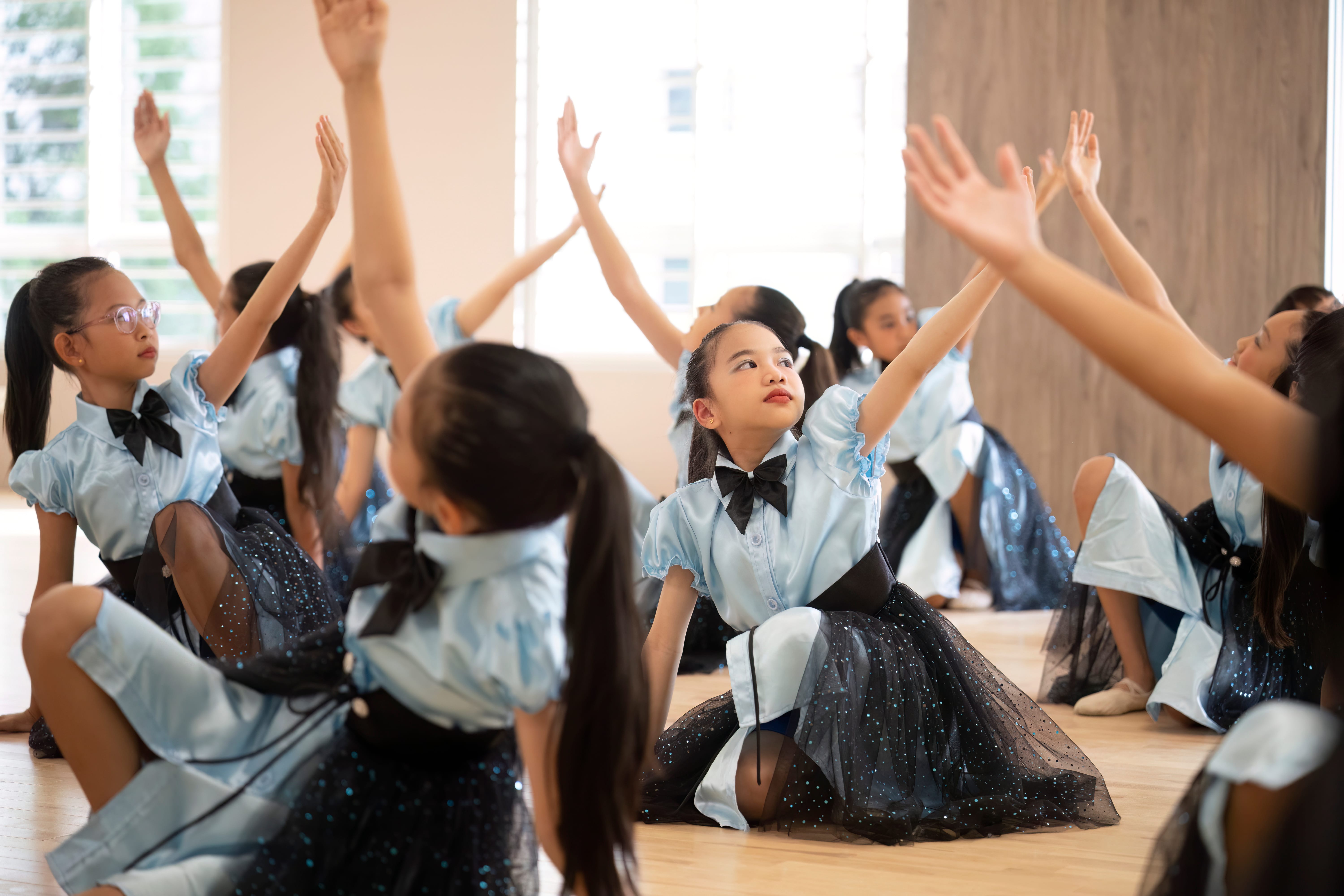 Girls in matching blue costumes kneel on a studio floor with arms raised in a dance pose.