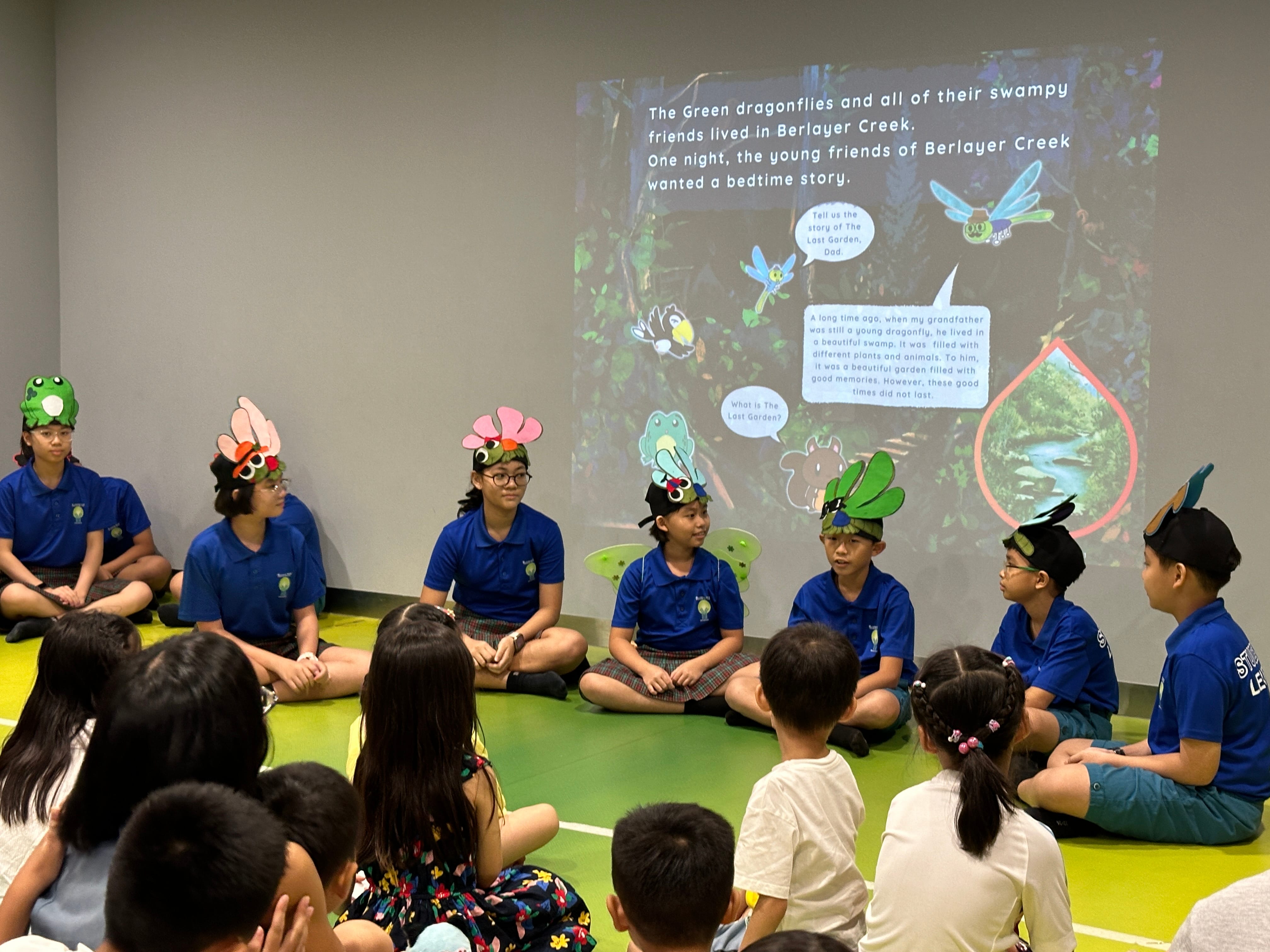 Children in blue uniforms wearing craft animal hats sit in a circle facing younger kids, with a storybook slideshow projected behind them.