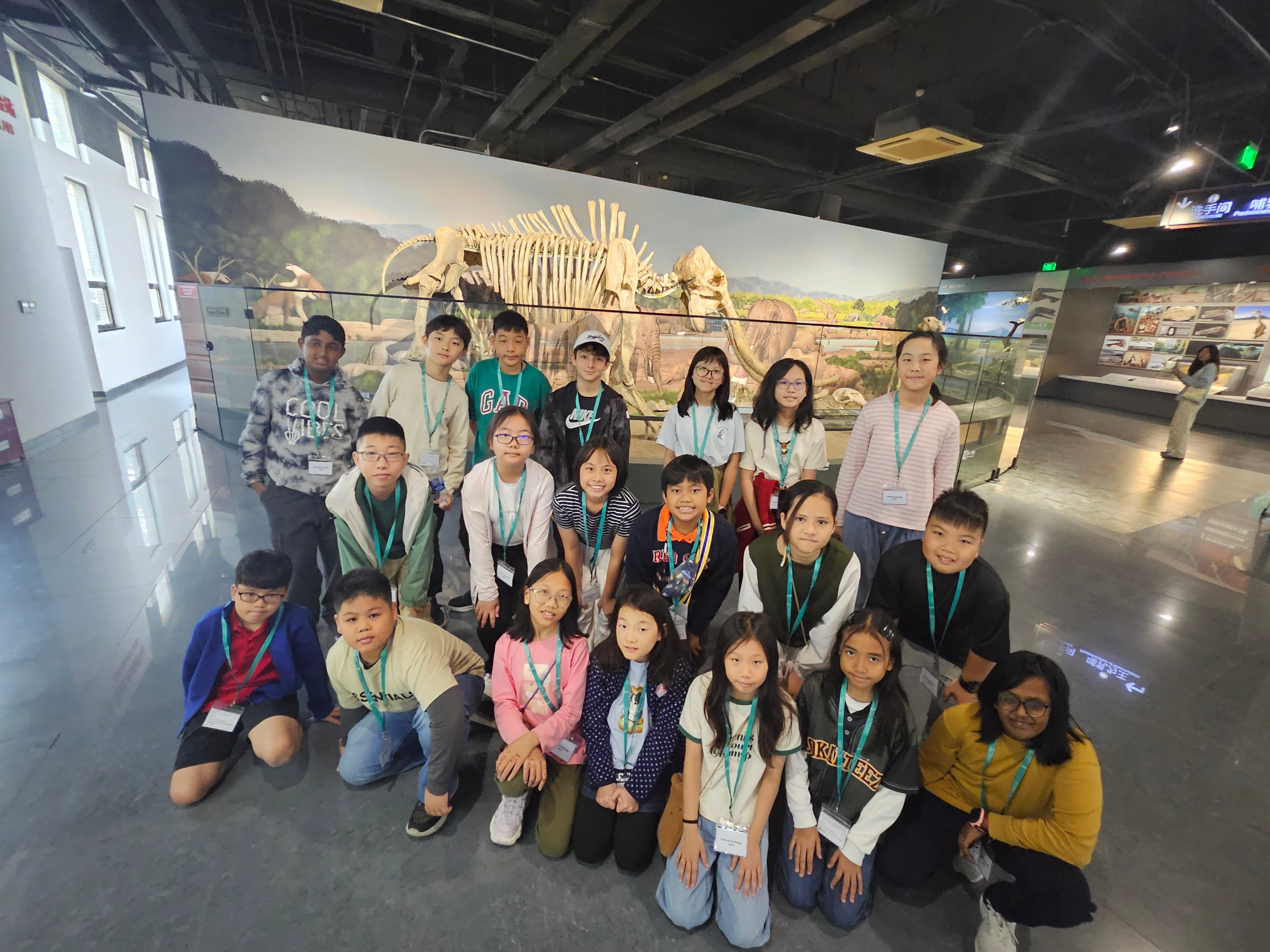 A group of about 20 children with lanyards pose in front of a large dinosaur skeleton exhibit in a museum.