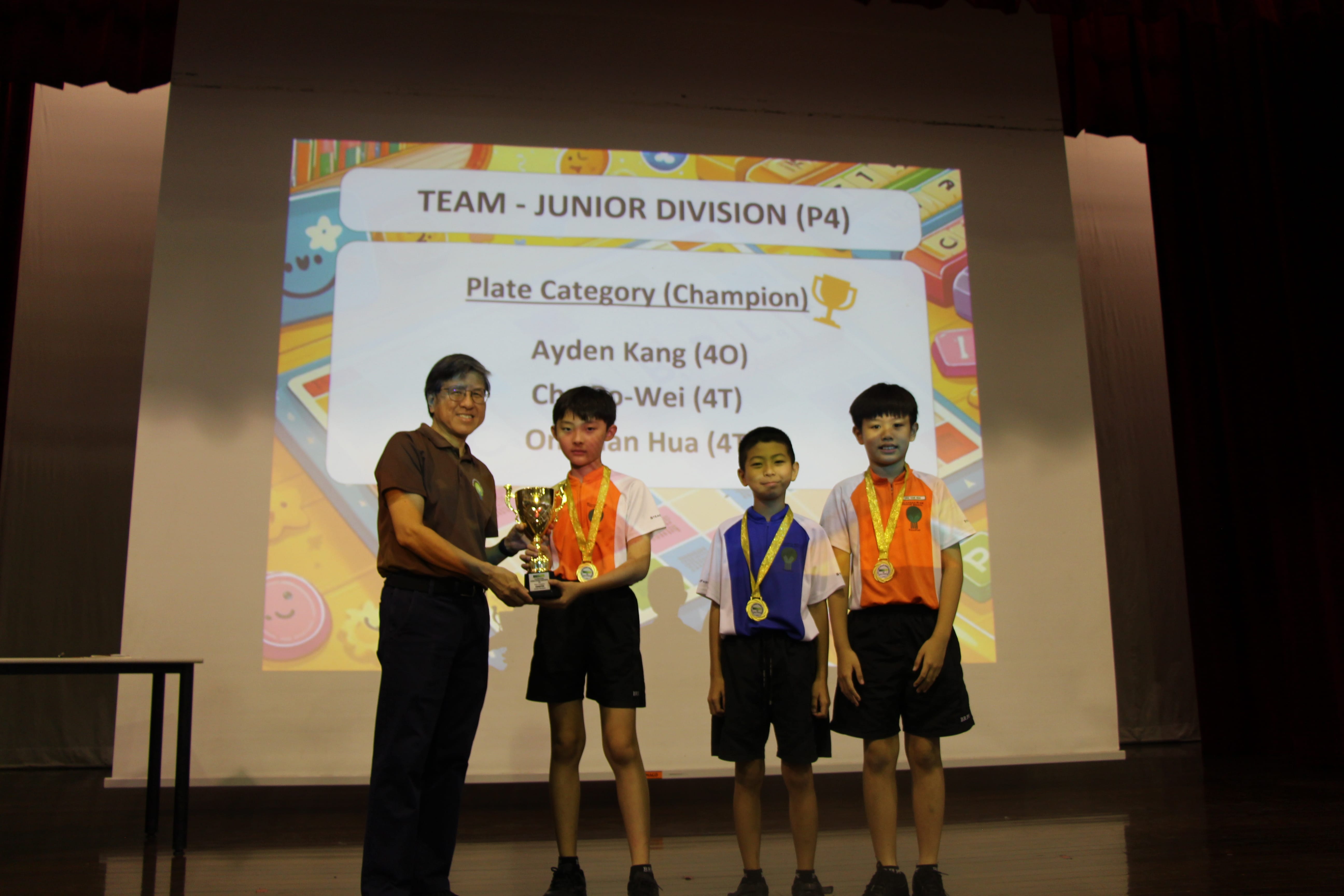 Man hands a trophy to a medal-wearing boy on stage; two other medal-wearing boys stand beside him.