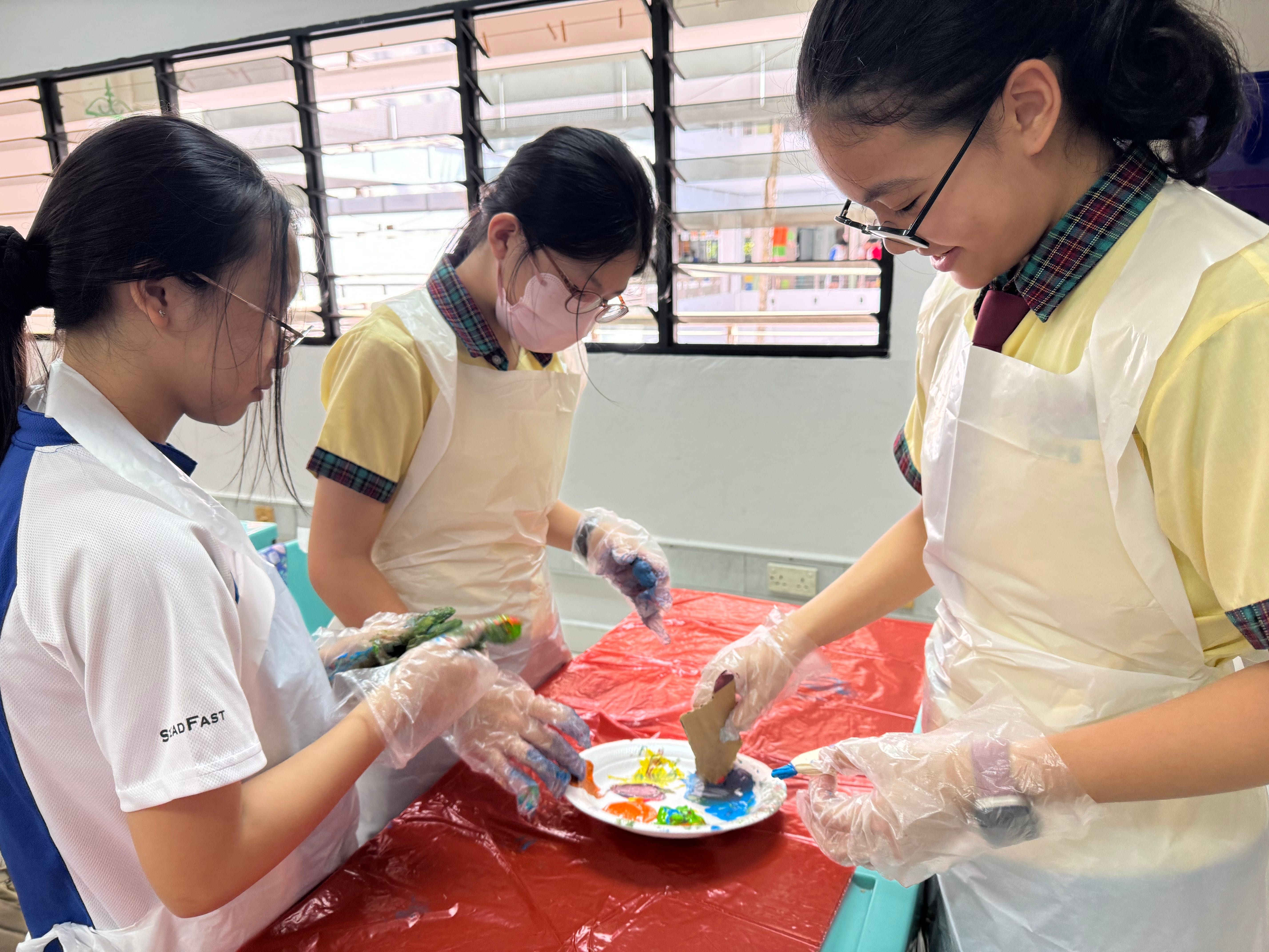 Three students in aprons and gloves mix paint on a paper plate at a red-covered table in a classroom.
