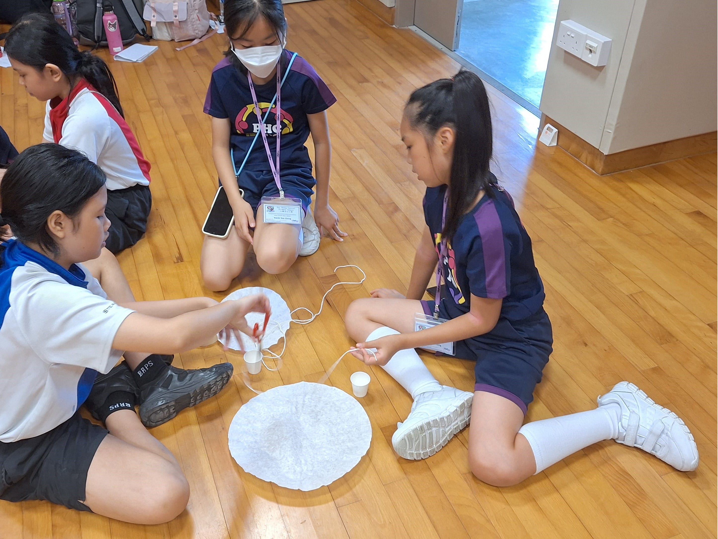 Three children sit on a wooden floor working on a craft project with white circular material, string, and small cups.