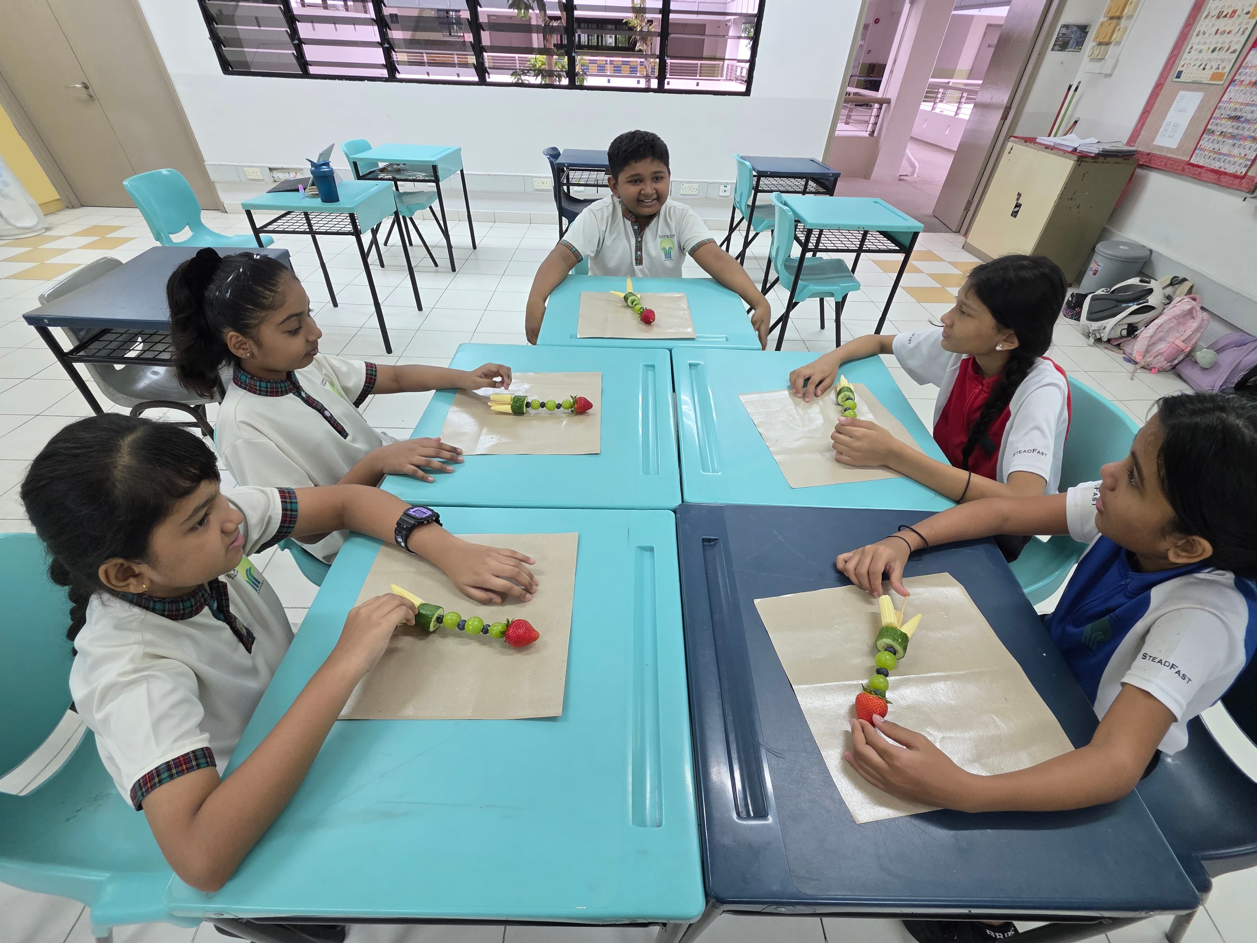 Five school children in uniforms sit around blue tables making fruit skewers with strawberries and kiwi.
