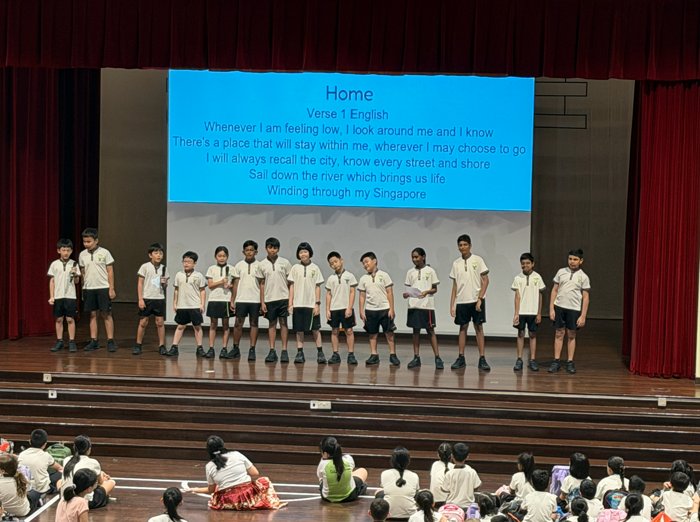 Students in uniform stand on a stage performing, with a screen showing lyrics to the Singapore song "Home."