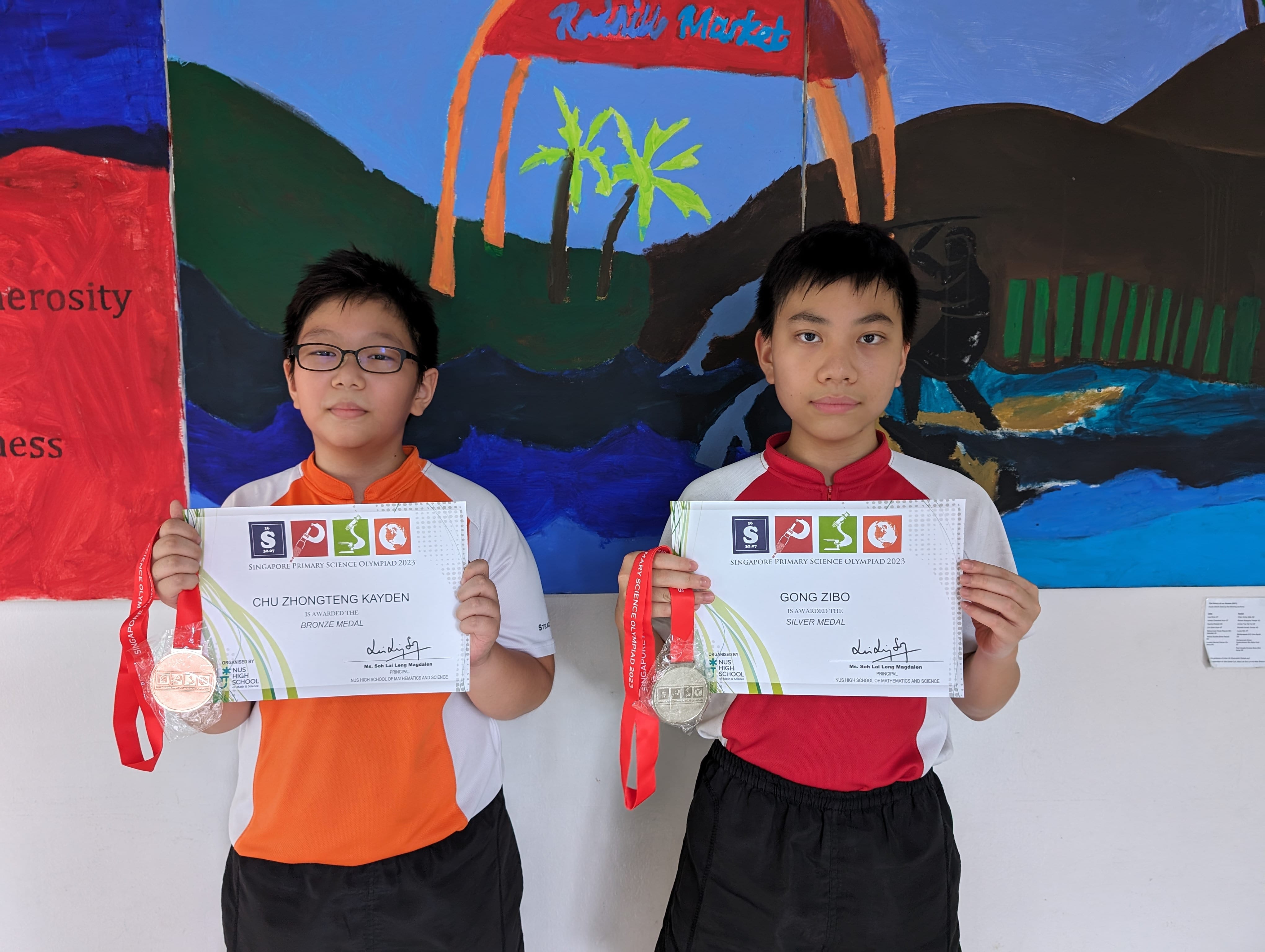 Two boys hold Singapore Primary Science Olympiad 2023 certificates and medals, one bronze and one silver.