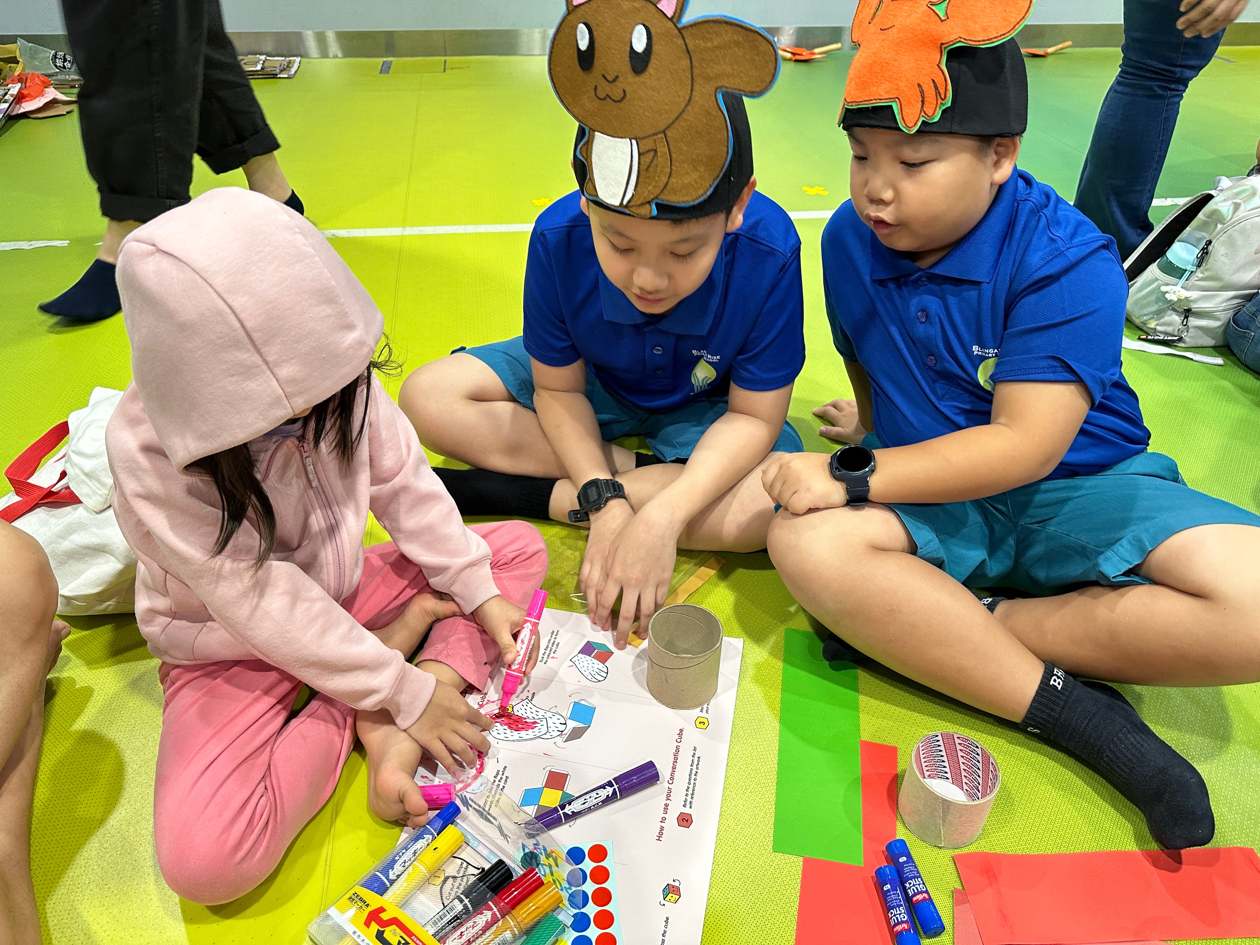 Three children sit on a green mat, coloring a worksheet; two boys wear animal hats and watch.