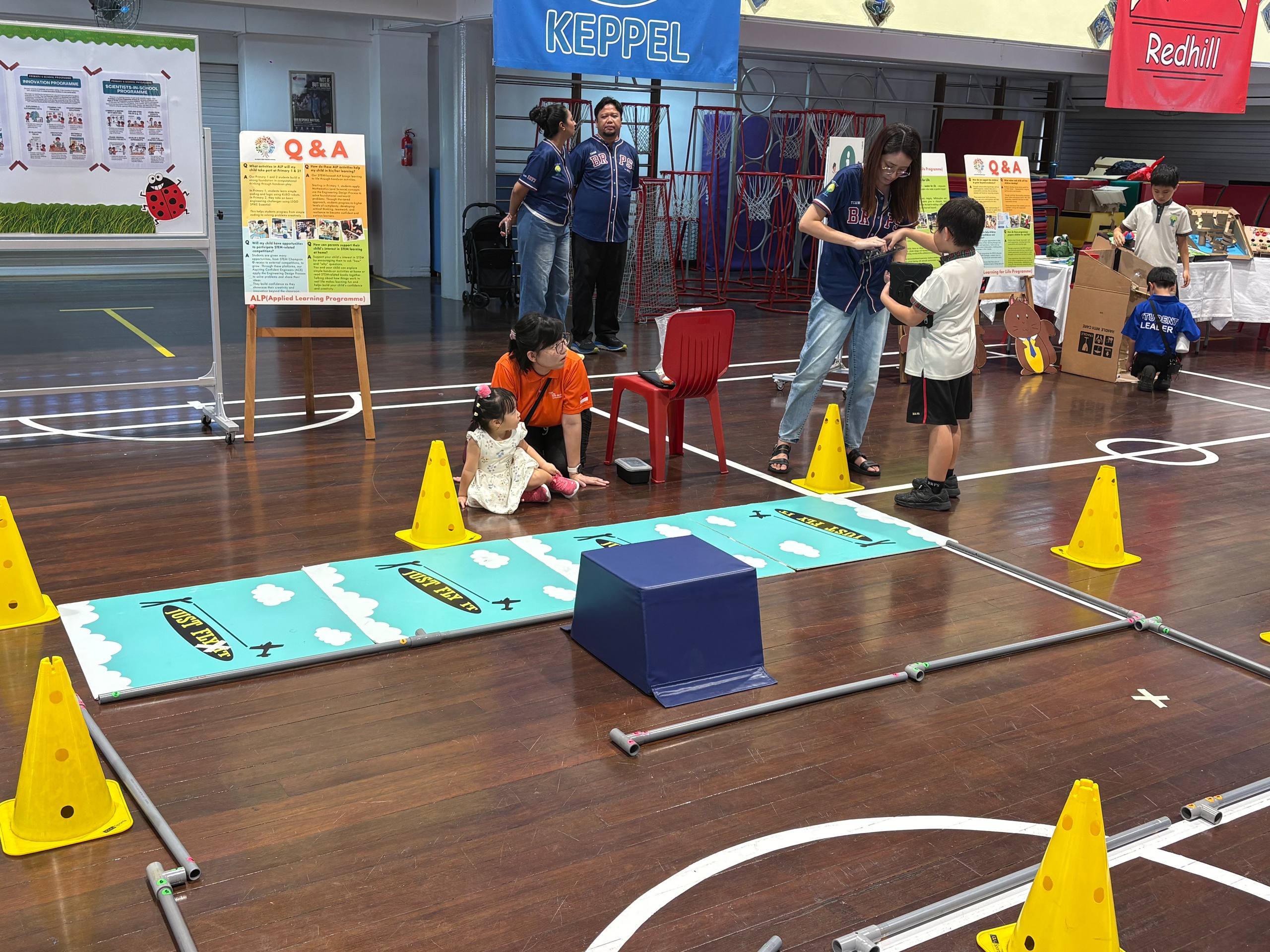Children and adults at a school activity day with yellow cones, mats, and Q&A boards in a gymnasium.