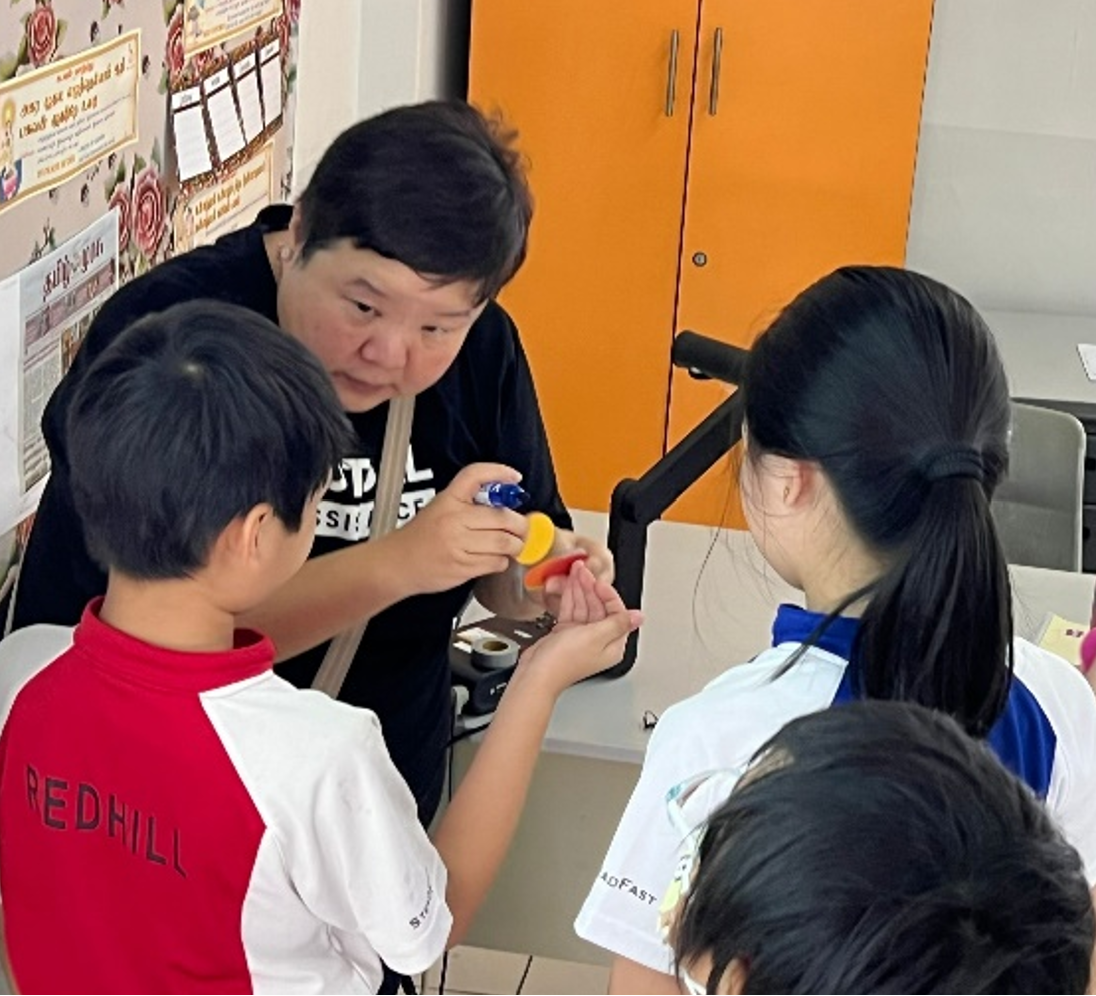 An adult in a black shirt leans over two children, demonstrating a hands-on activity with small colorful objects.