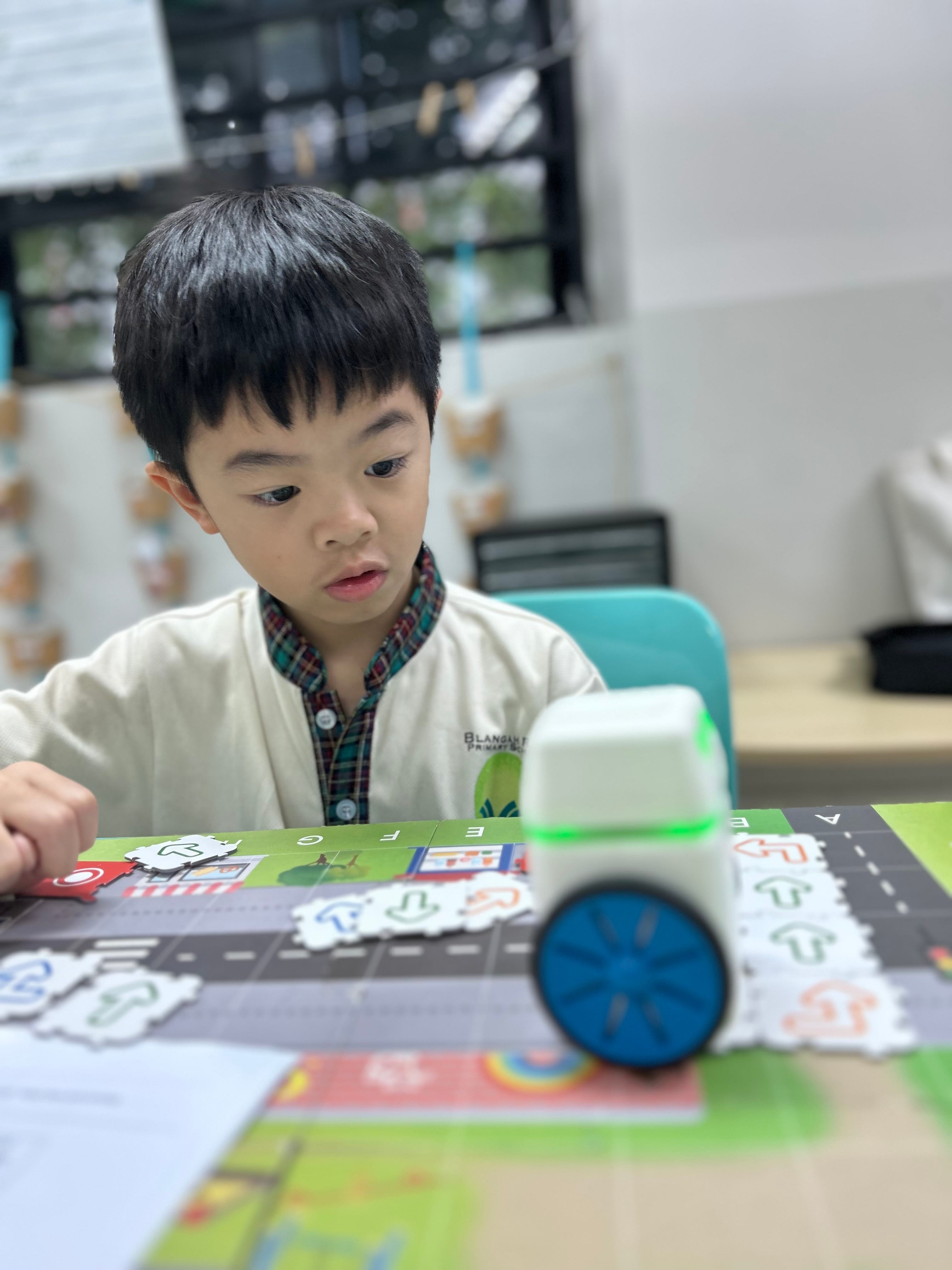 Young boy in school uniform focuses on a small wheeled robot with a green light on a colorful activity mat.