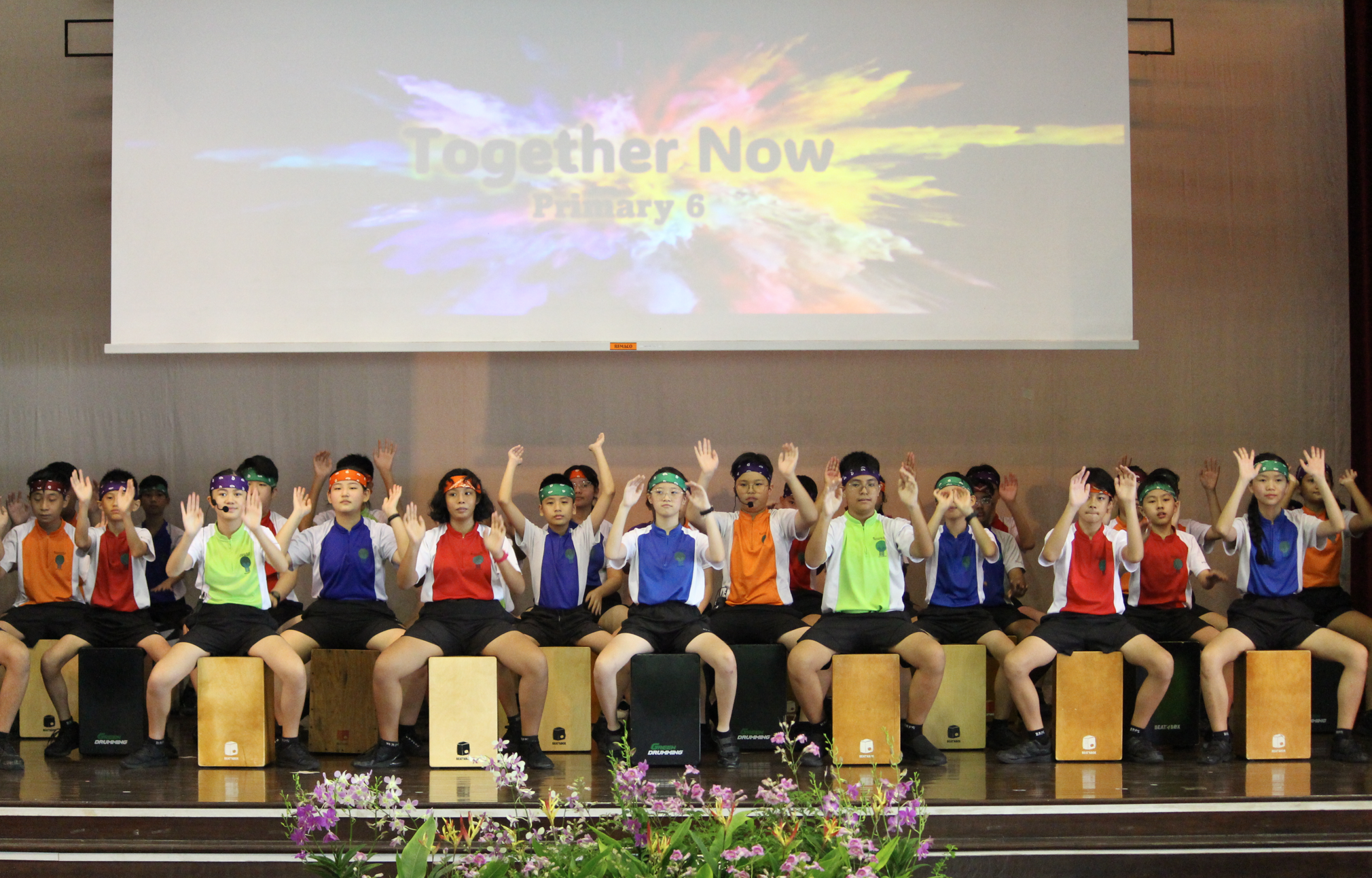 Children seated on cajón drums on stage, hands raised; projection reads Together Now Primary 6.