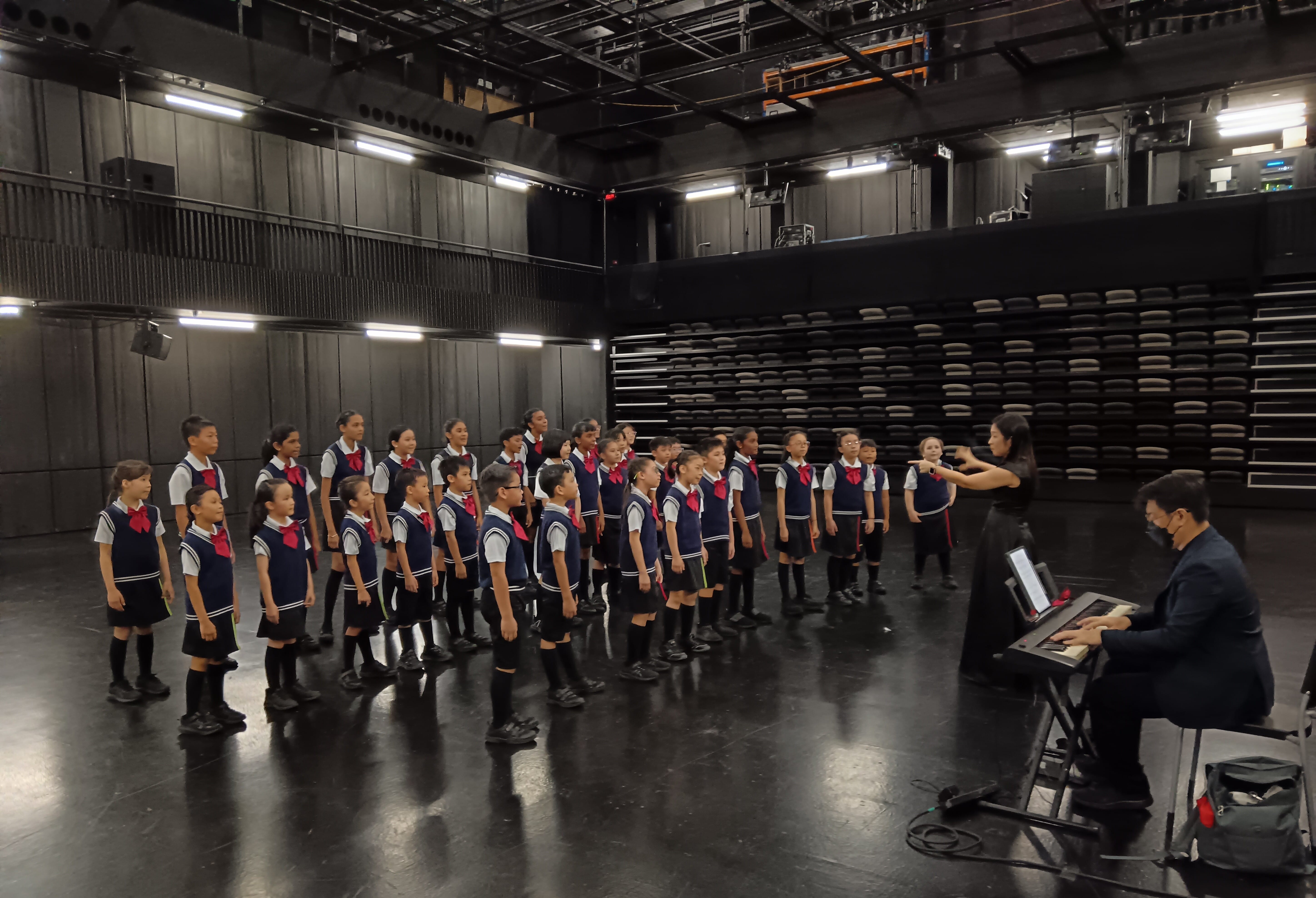 A children's choir in navy uniforms with red ties rehearses on a black stage, led by a conductor and a keyboard player.
