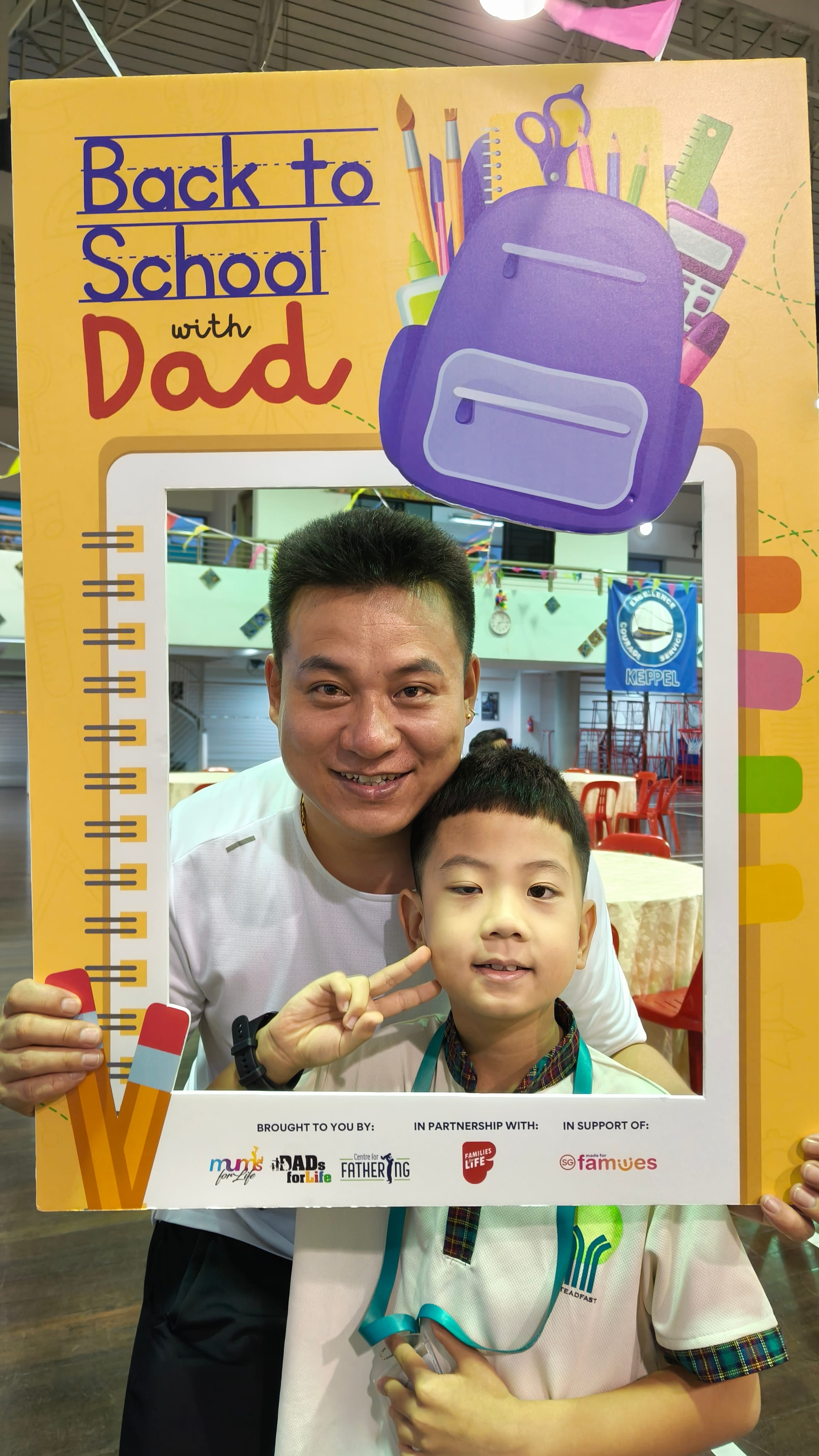 A man and young boy smile behind a colorful "Back to School with Dad" photo frame at an indoor event.