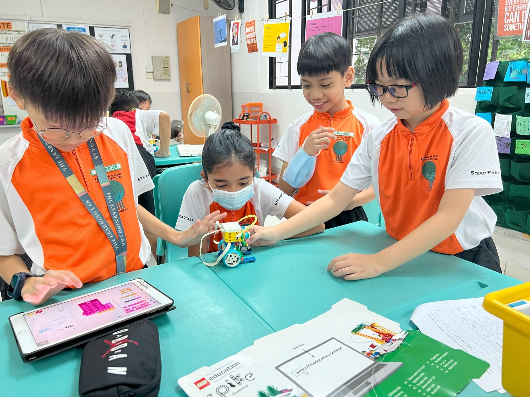 Four children in orange school uniforms work together on a small LEGO robot at a teal table, using a tablet to program it.