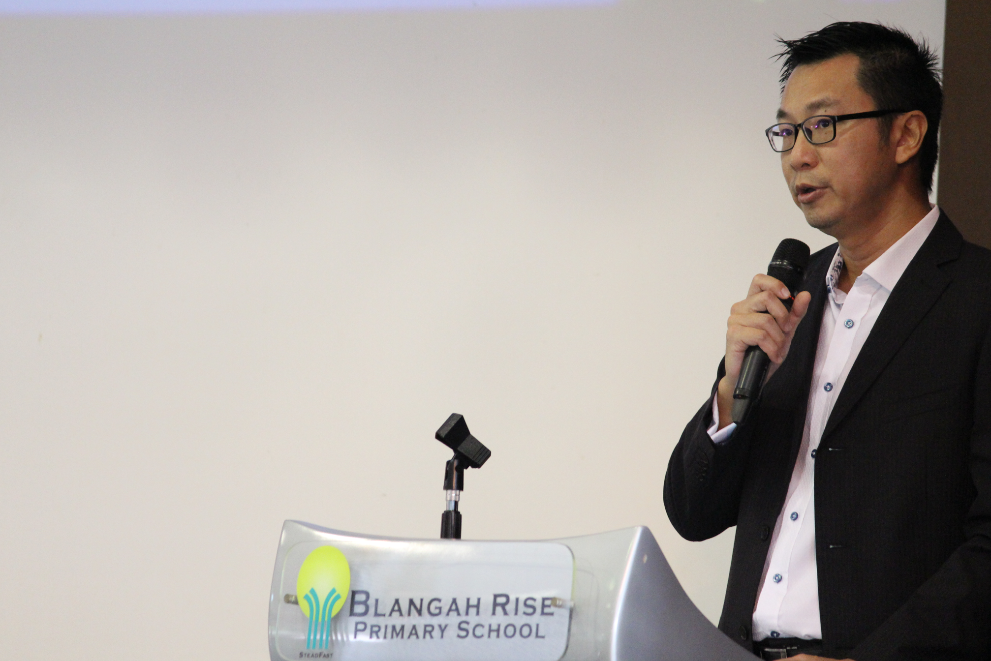 Man in glasses and black blazer speaking into a microphone at a Blangah Rise Primary School podium.