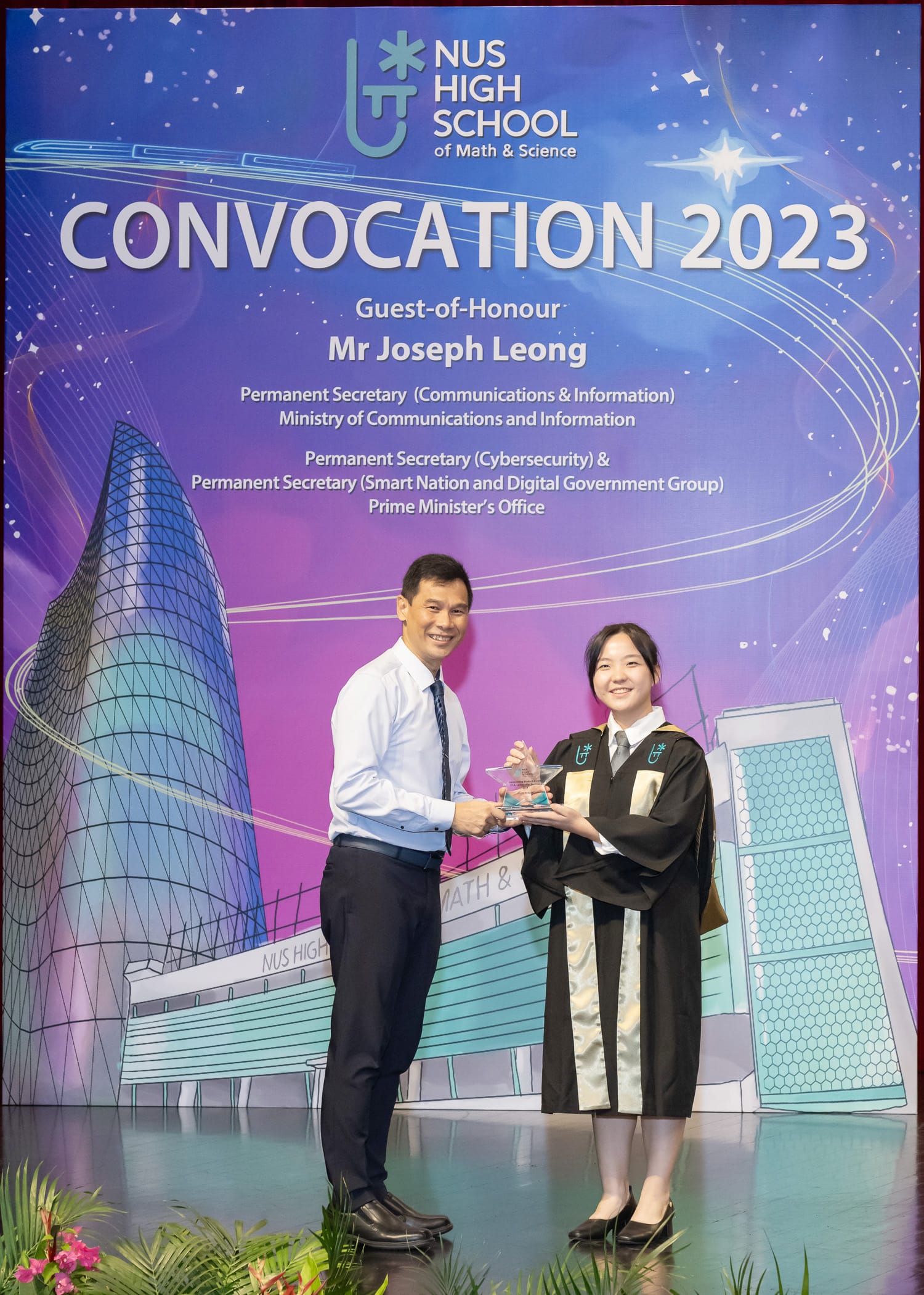 A man and a graduating student in black robes hold a trophy at NUS High School Convocation 2023 backdrop.
