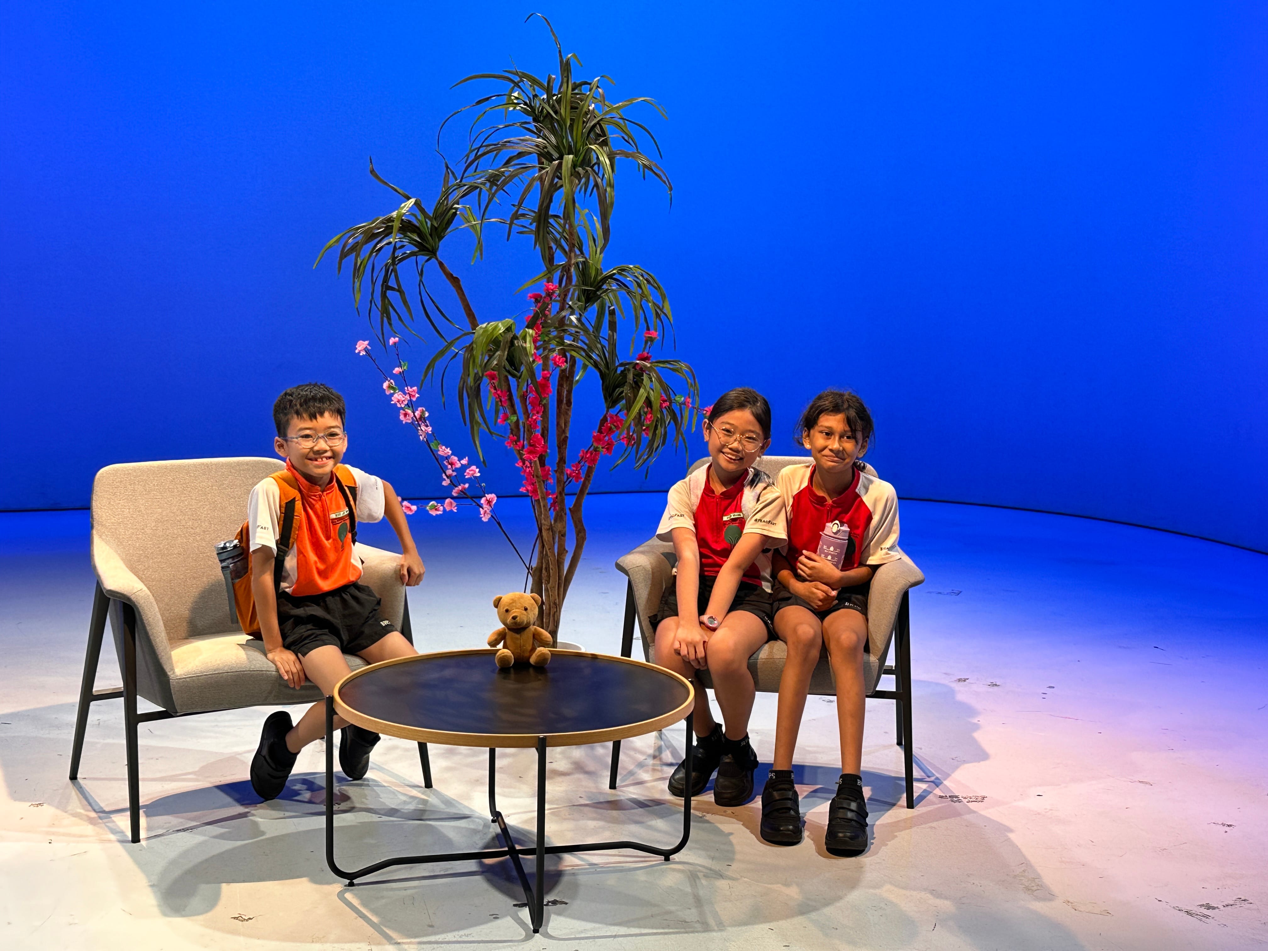 Three children in school uniforms sit on chairs around a small round table with a teddy bear, in front of a blue backdrop.