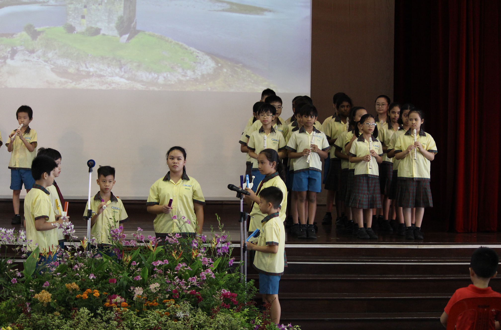 Schoolchildren in yellow uniforms stand on stage holding recorders near microphones, flowers in front.