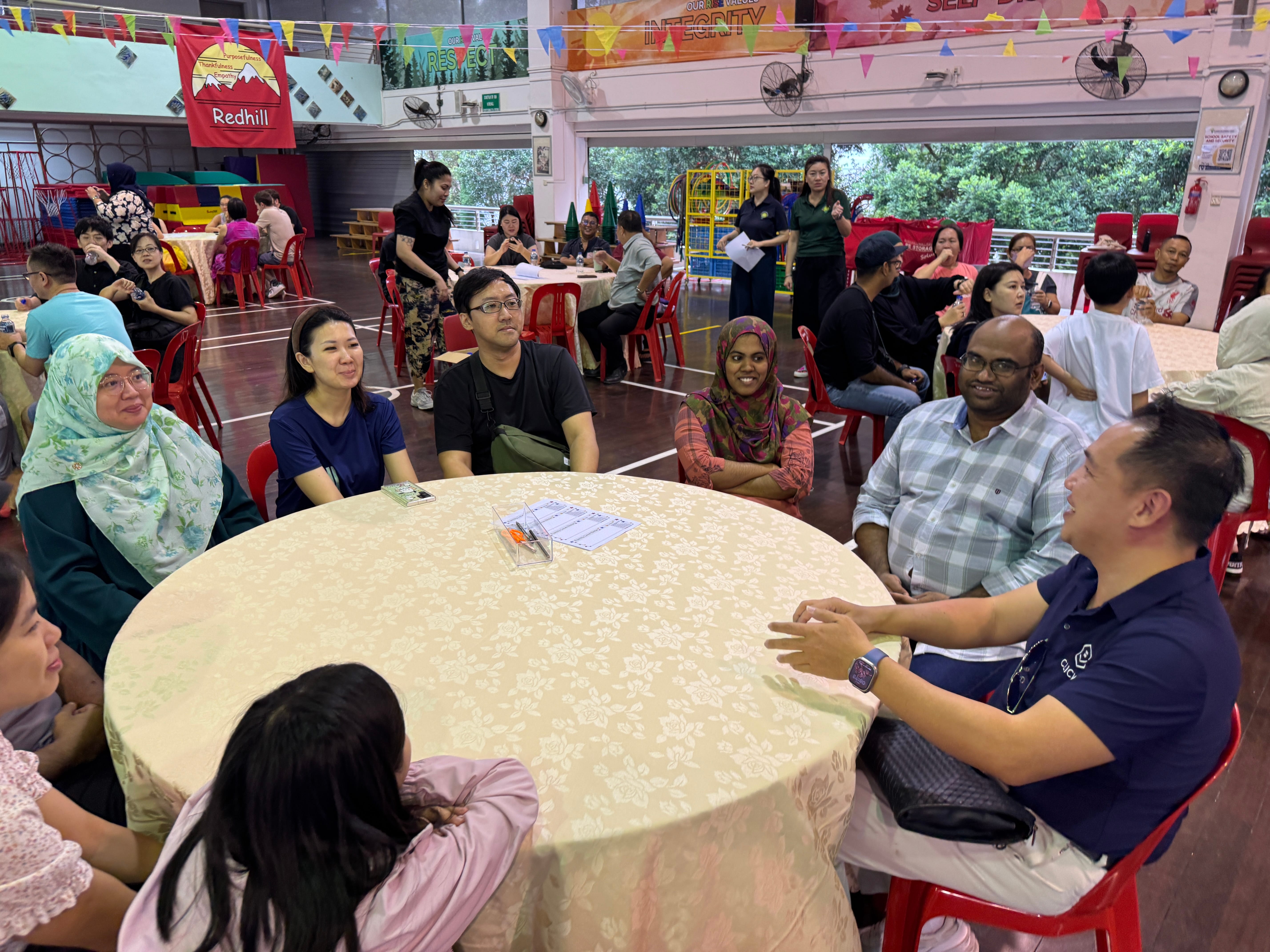People seated around round tables in a hall with bunting overhead; group chatting at a large table.