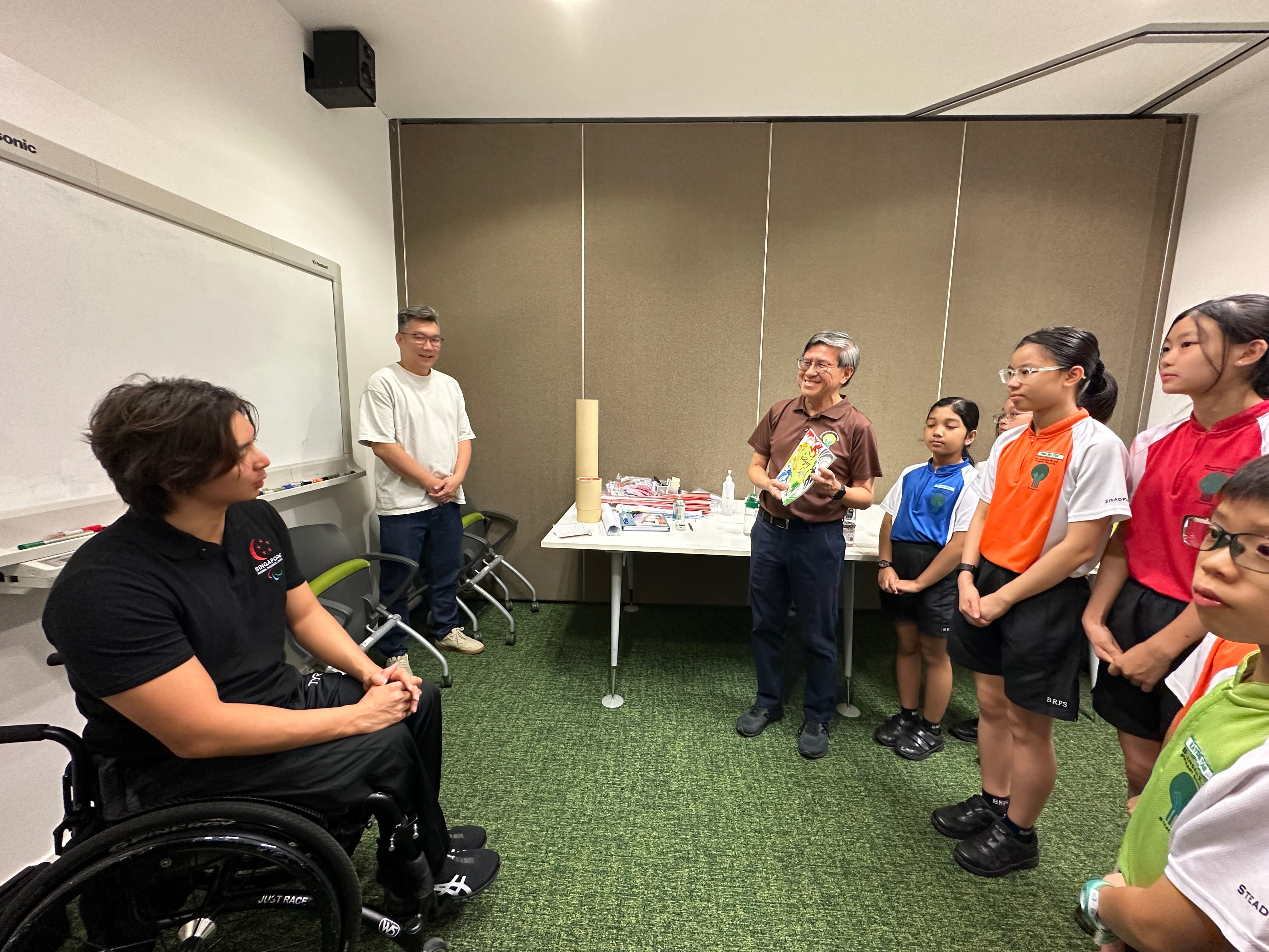 A man in a wheelchair faces an older man holding a book, surrounded by schoolchildren in a classroom.