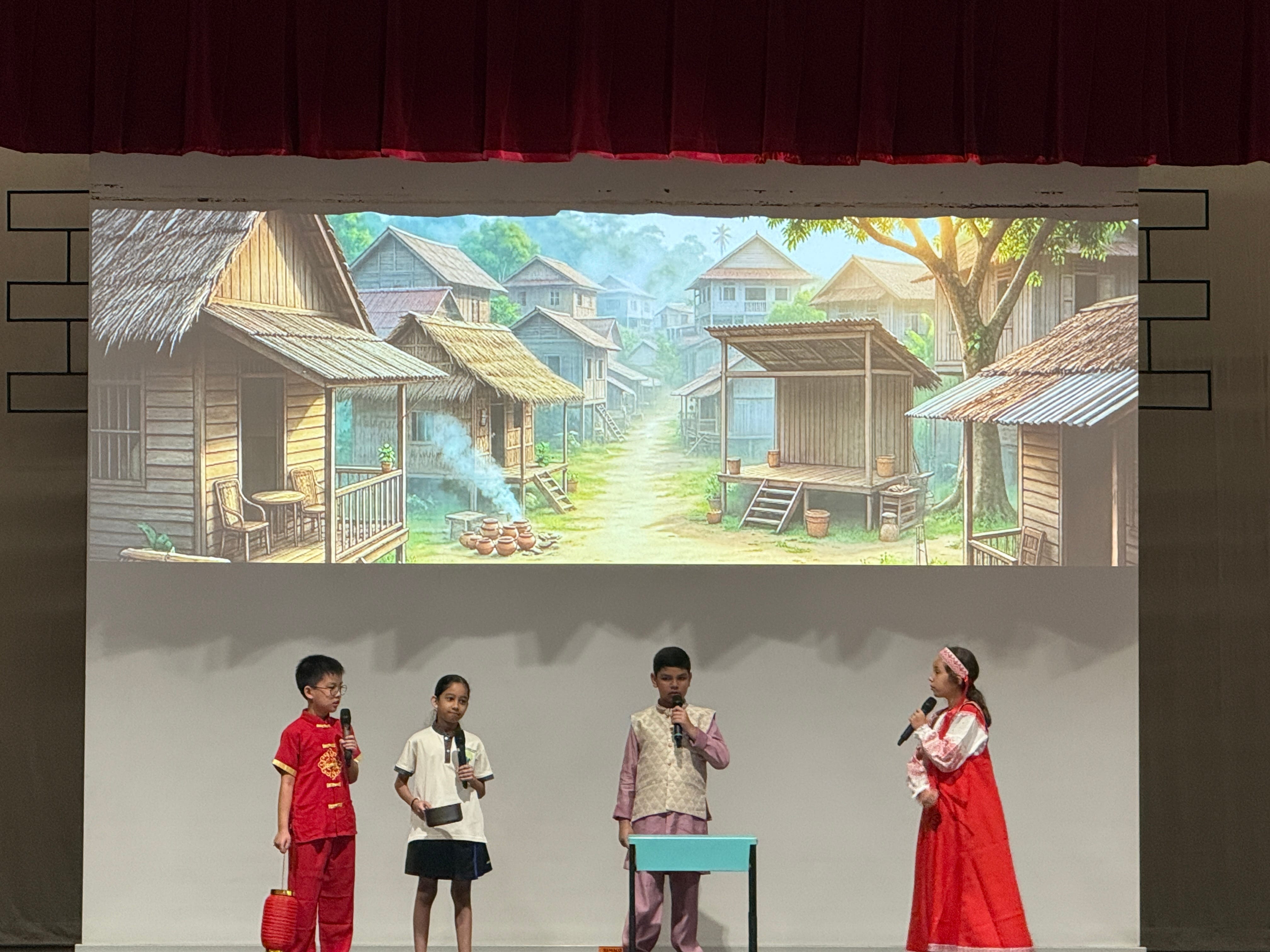 Four children in cultural costumes hold microphones on a stage with a projected village scene backdrop.