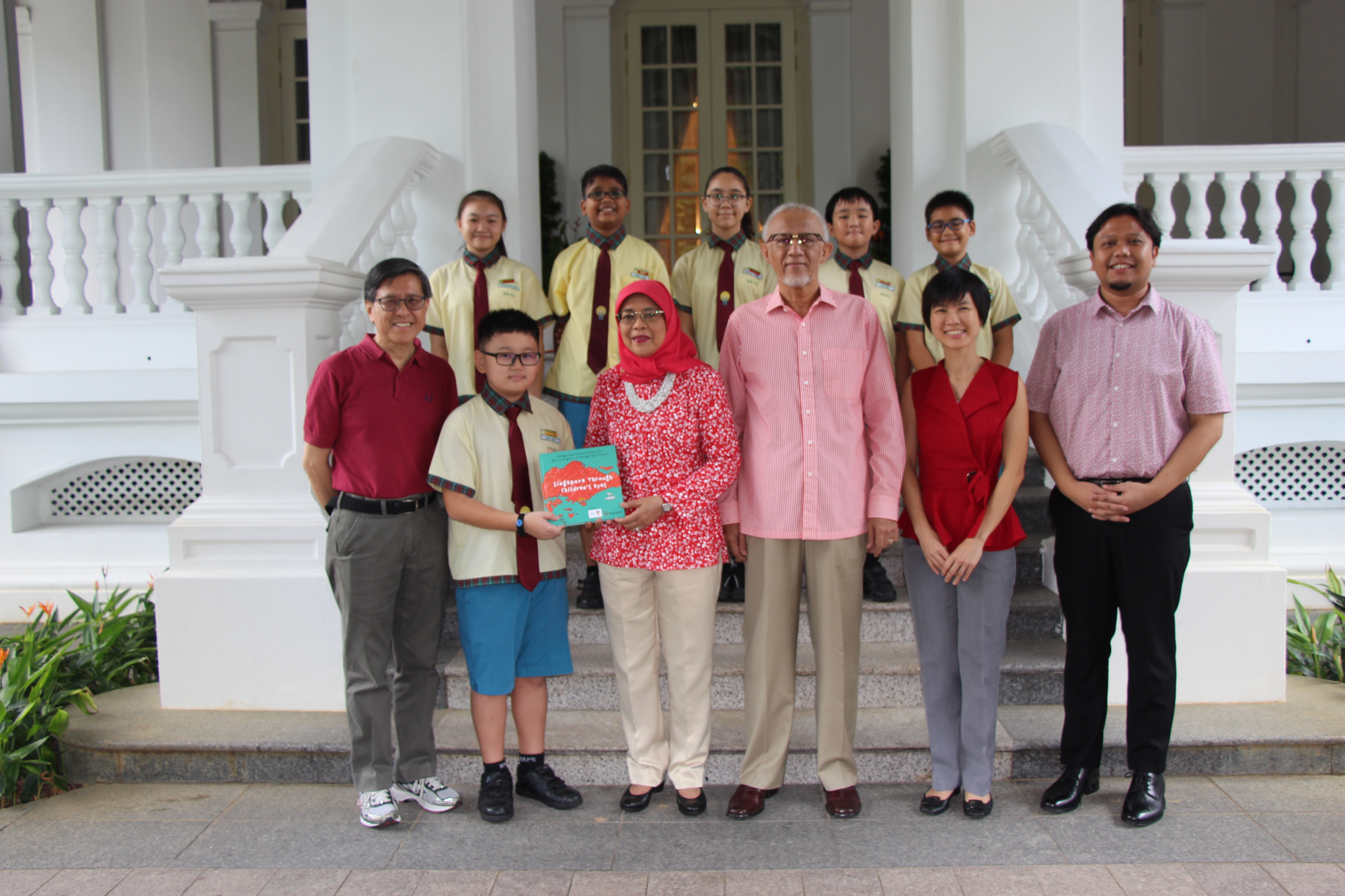 A group of adults and children in school uniforms pose on steps of a white colonial building, holding a teal book.