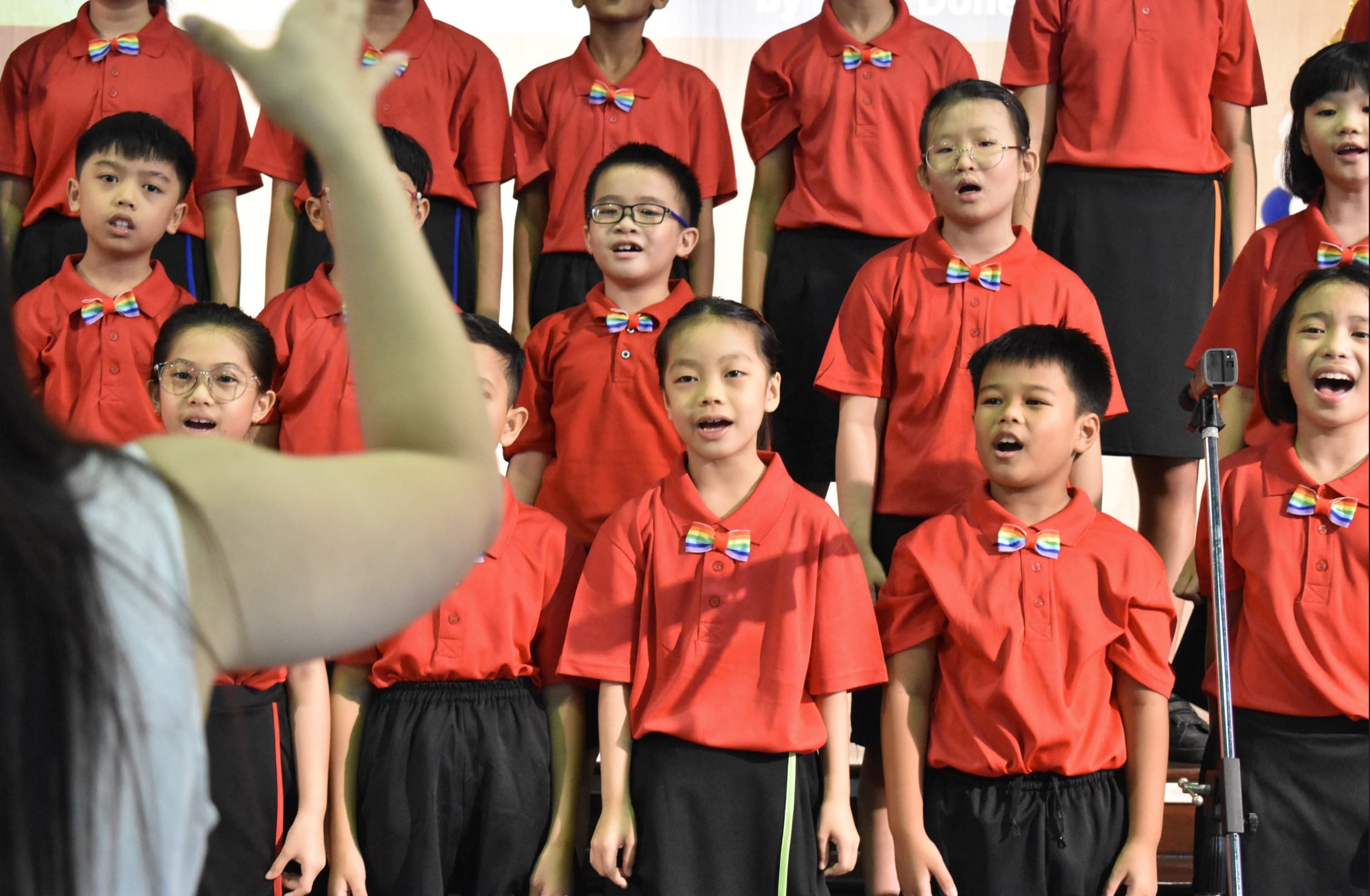Children in red shirts and rainbow bow ties singing in a choir, led by a conductor's raised hand.