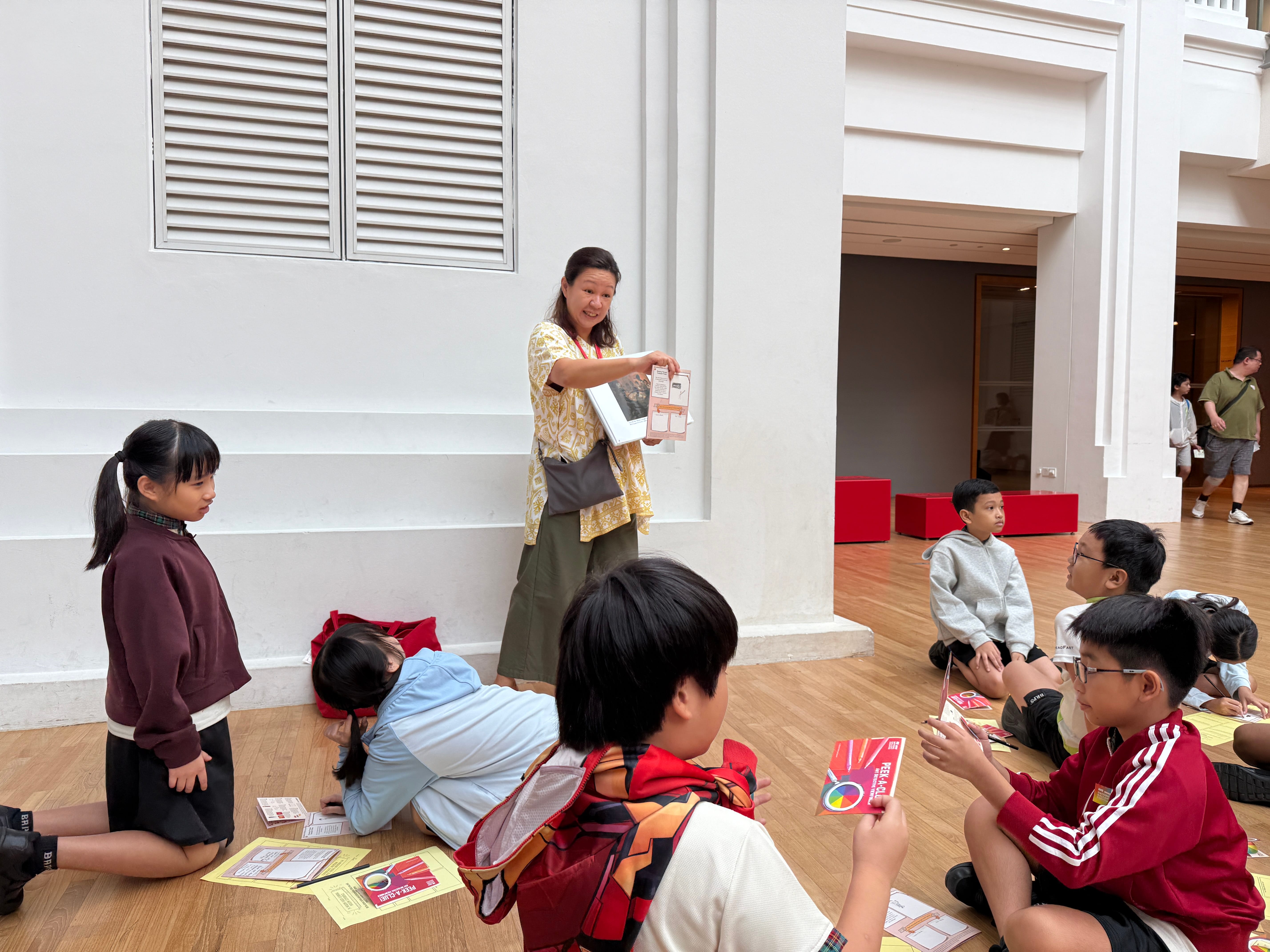 Woman holds up a booklet while children sit on a wooden floor with papers in a bright hall.