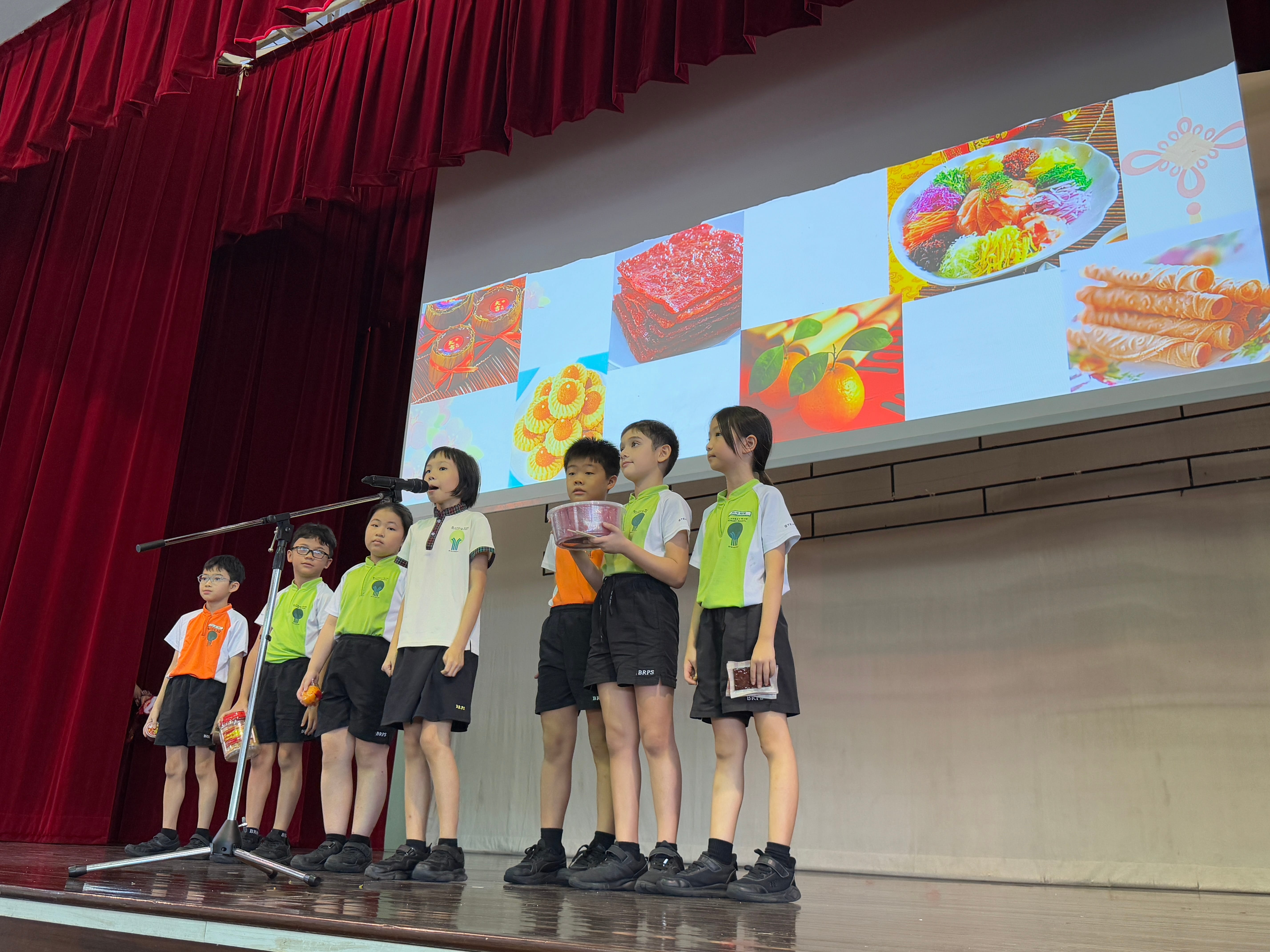 Seven children on a stage by a microphone, with a screen behind showing photos of colorful foods.