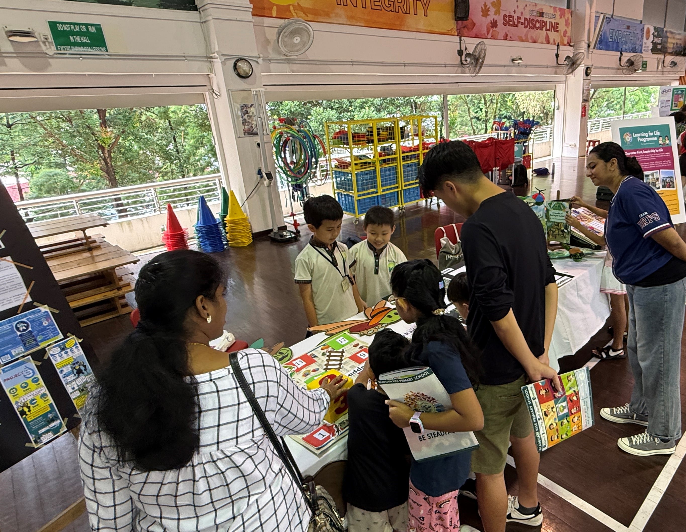 Adults and children gathered around a table game in a bright hall with large windows and posters overhead.