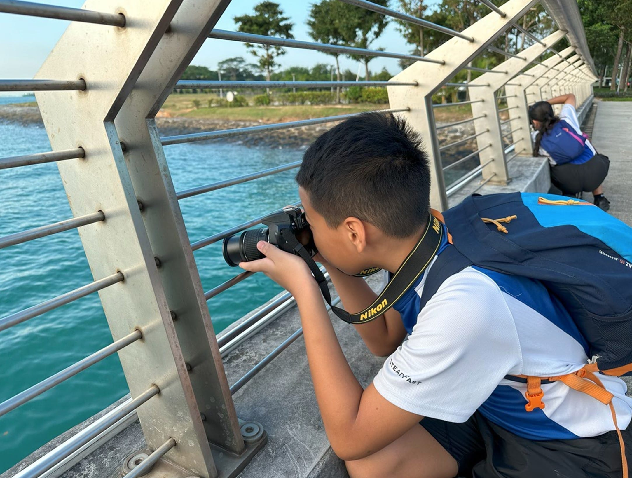 A boy with a backpack photographs through bridge railings over blue water, with another person crouching behind him.