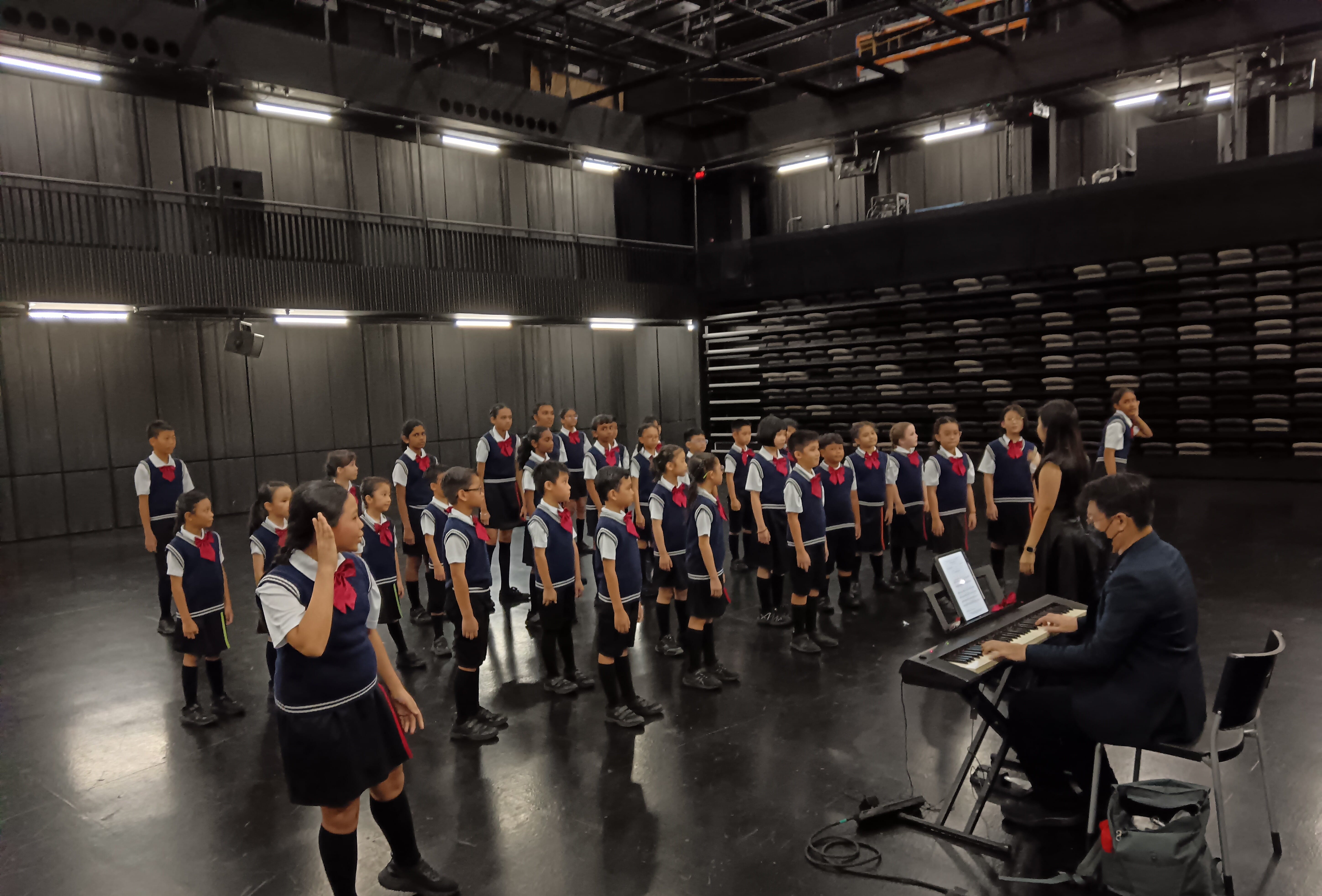 Children in navy uniforms with red ties rehearse as a choir in a dark studio while a person plays a keyboard.