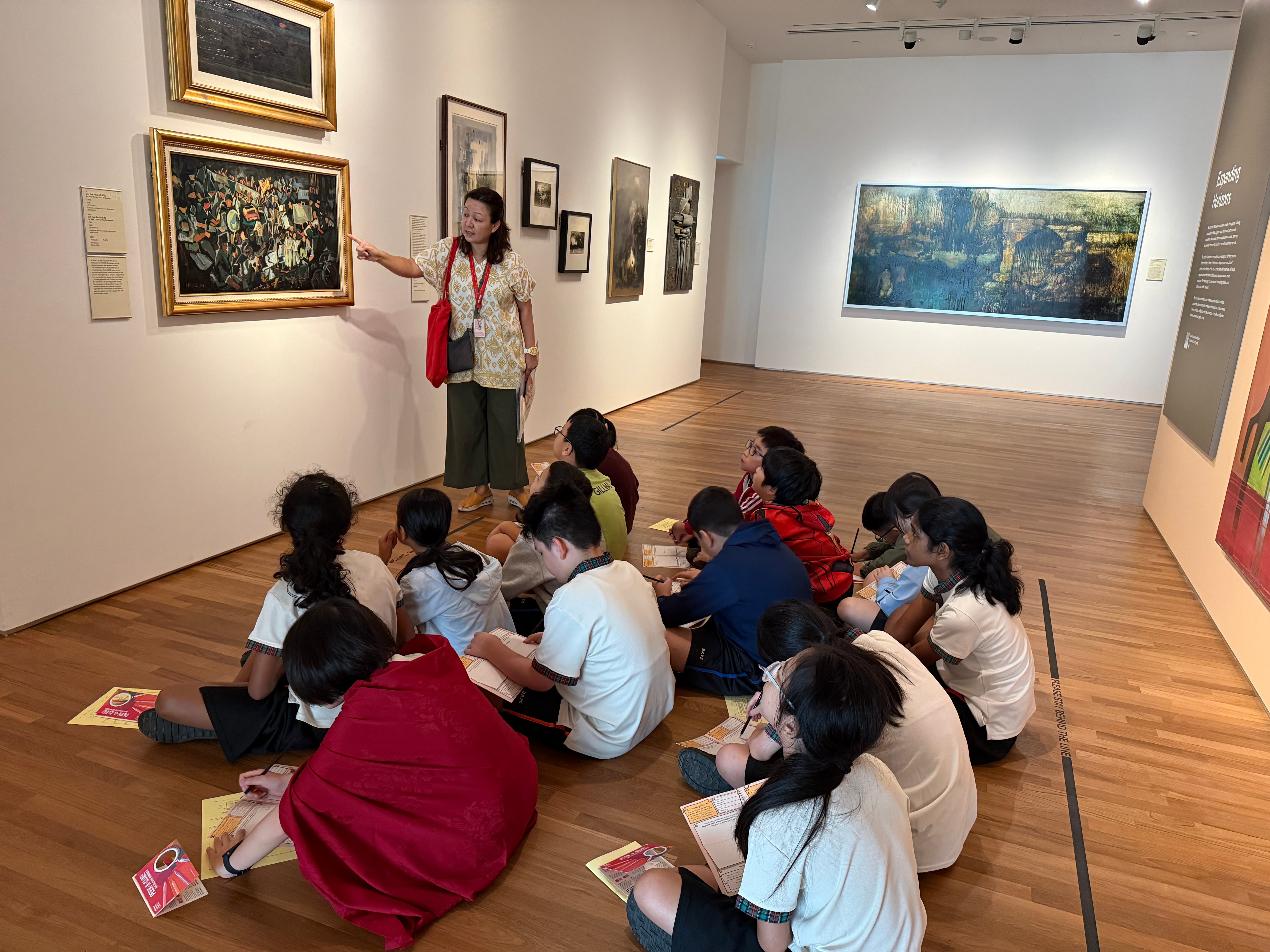 Teacher points at a framed painting in a museum gallery while seated students take notes on the floor.