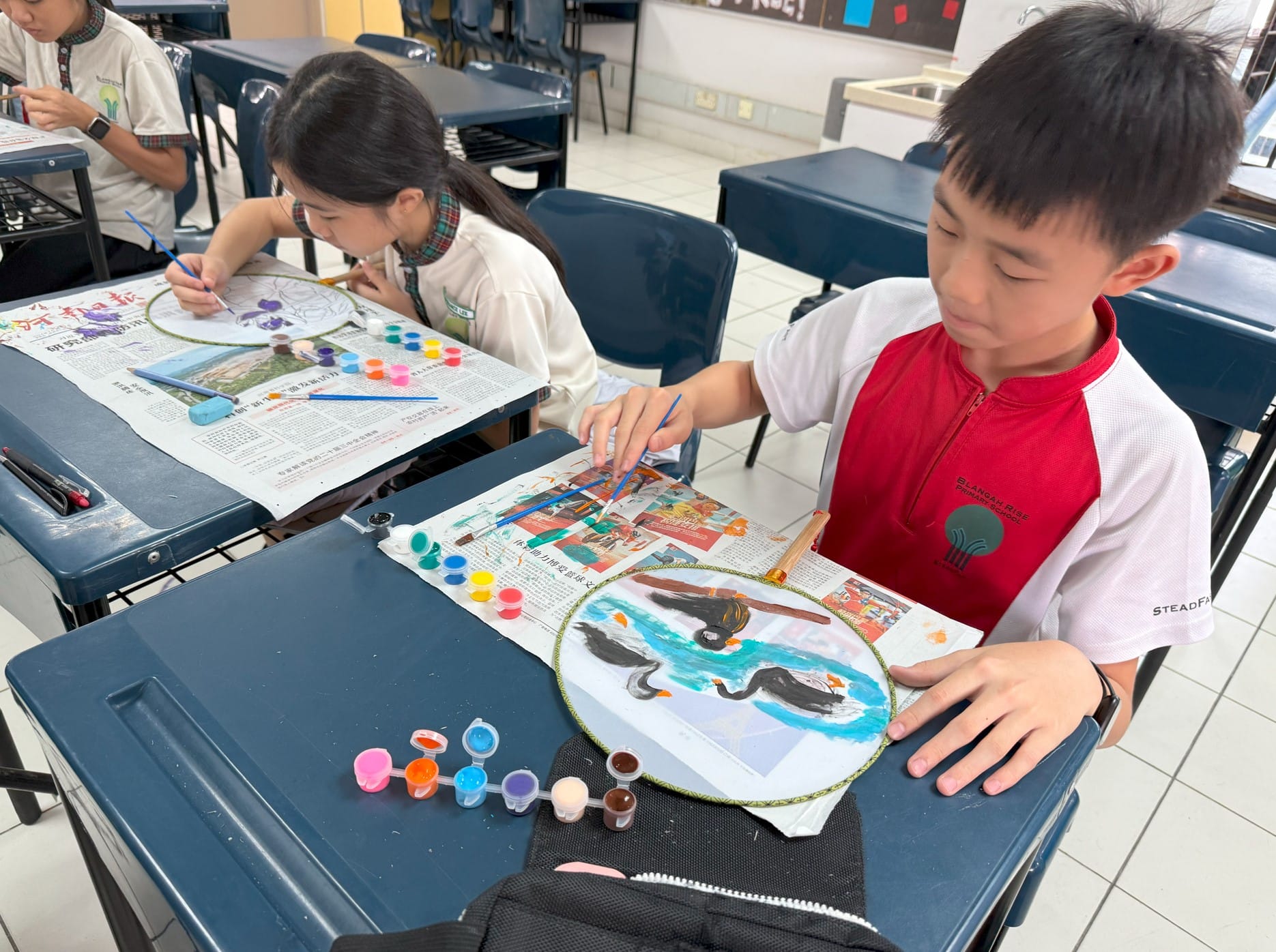 Two school children paint circular artworks with colorful paints on newspaper-covered desks in a classroom.