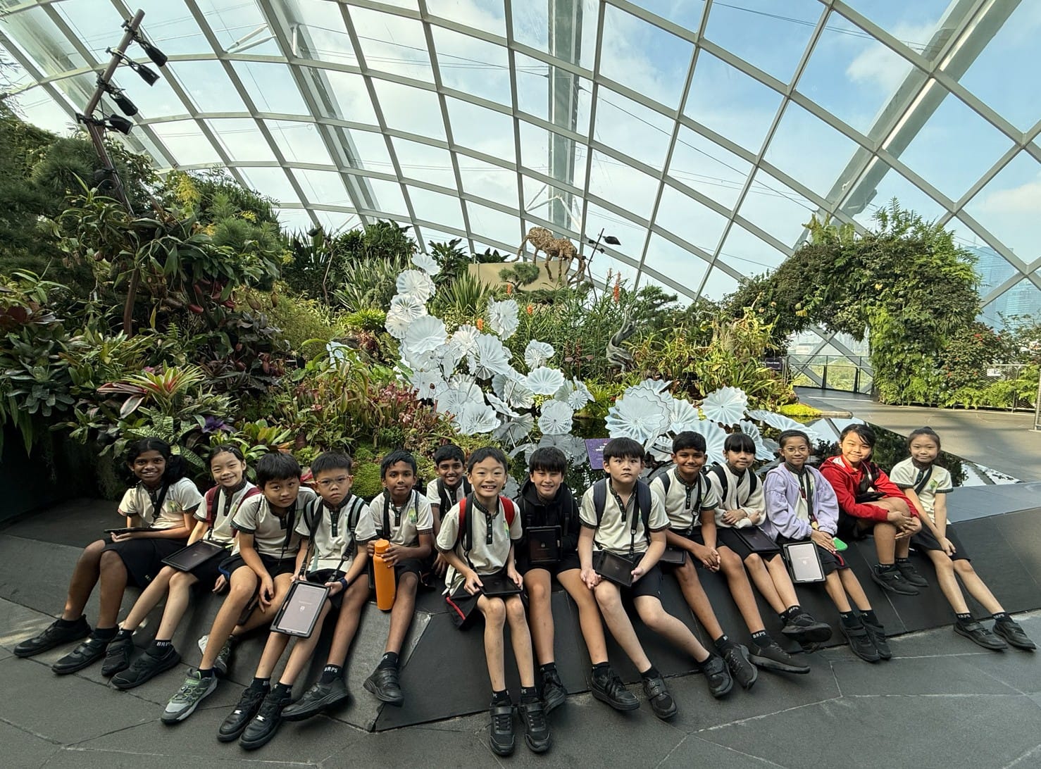 A group of uniformed school children sit together inside a large glass greenhouse filled with tropical plants.