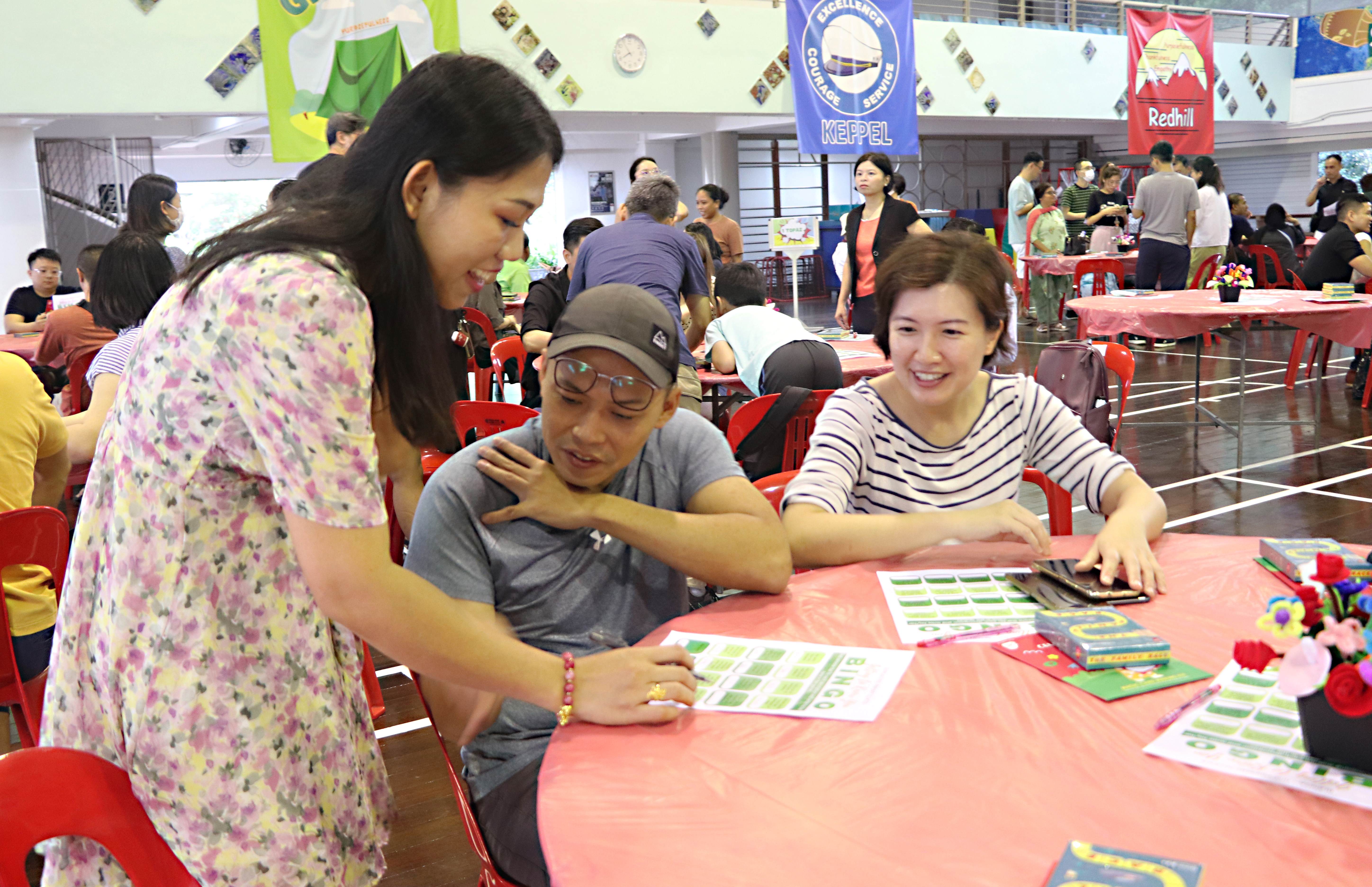 Three smiling people gathered around a pink-covered table playing bingo in a busy hall with Keppel and Redhill banners.
