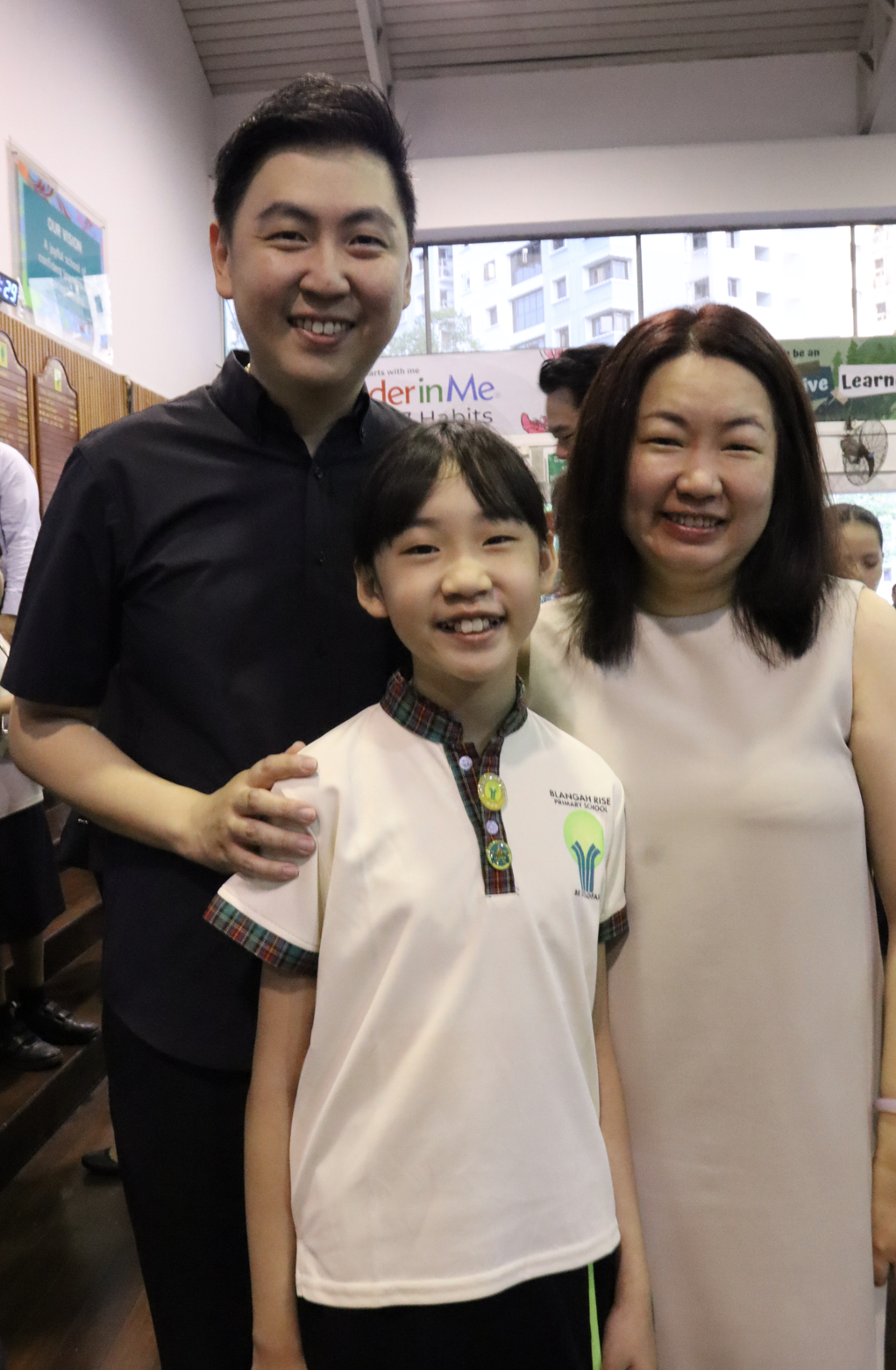 Two smiling adults pose with a child wearing a Blangah Rise Primary School uniform indoors.
