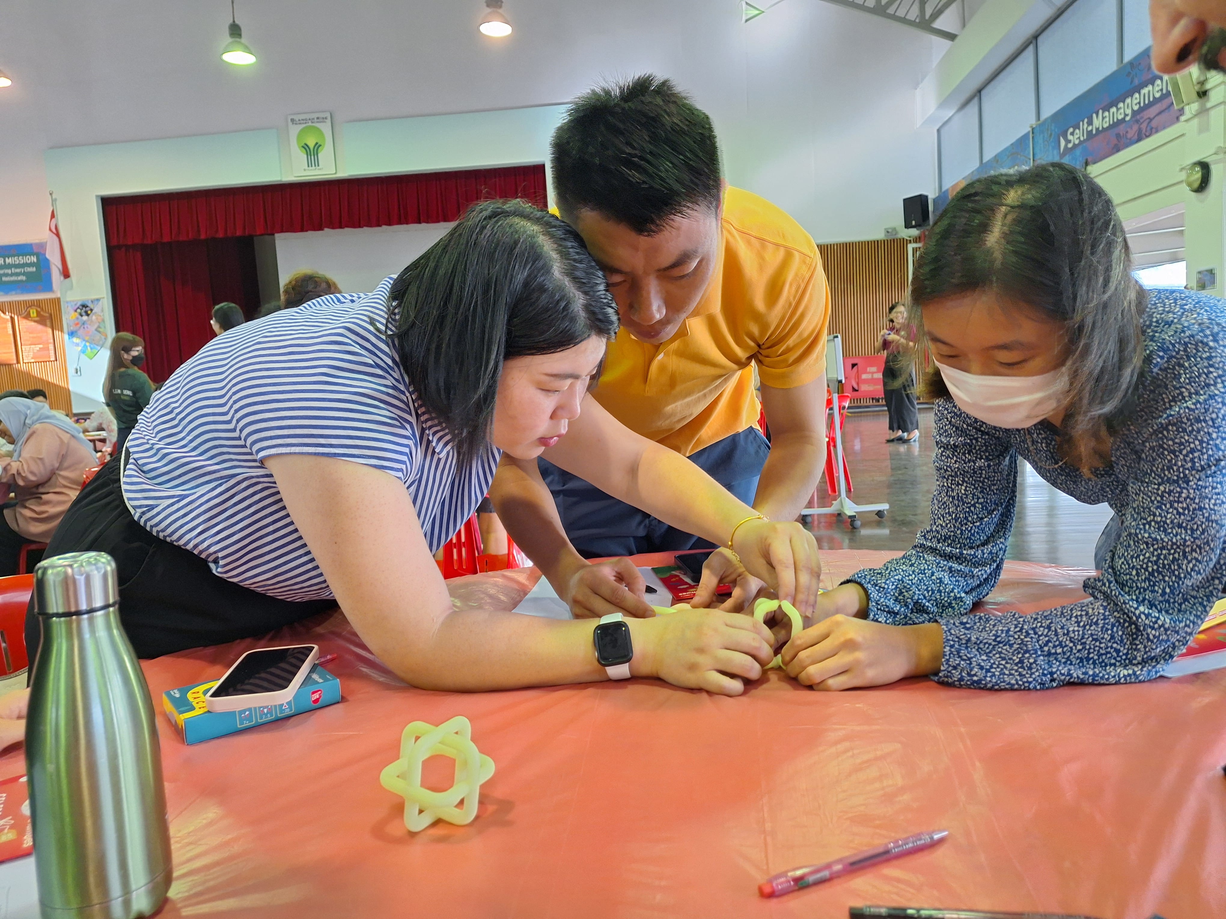 Three people lean over a red-covered table, working together with small linked pieces.