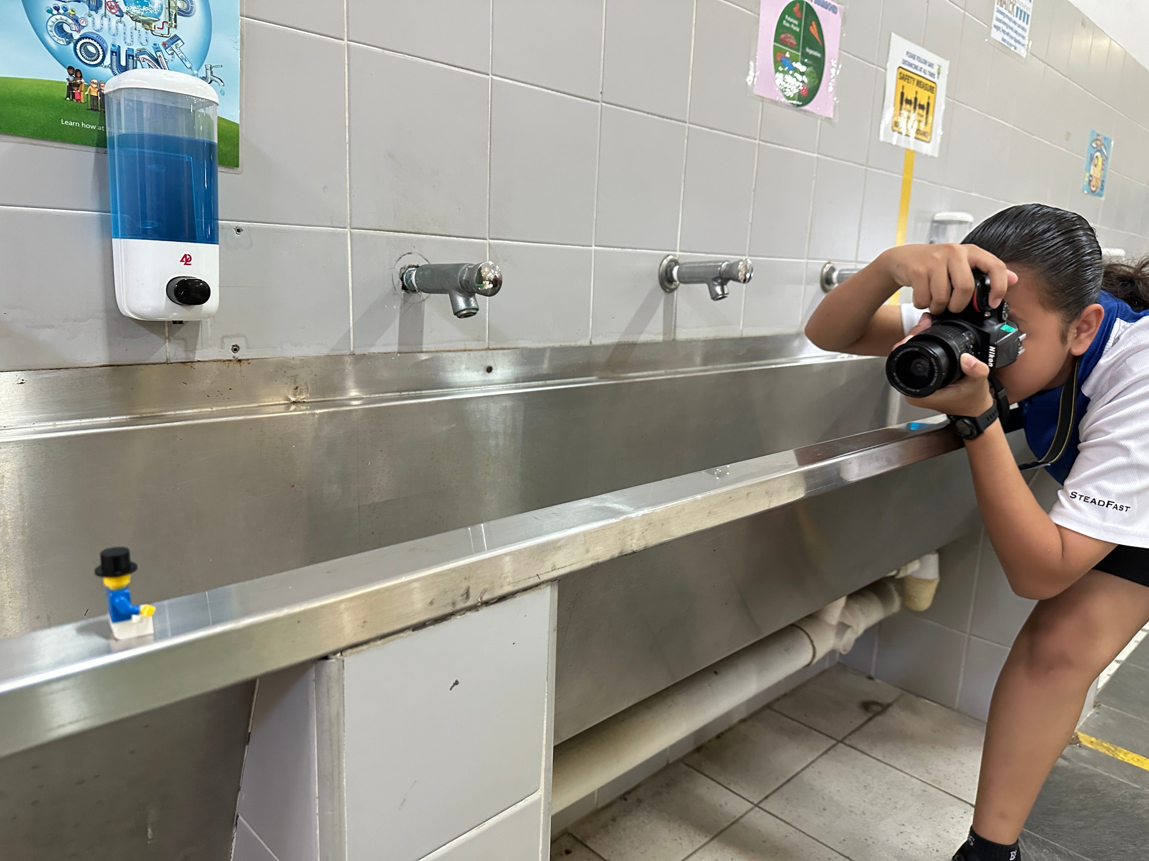 A child photographs a small Lego figure on a stainless steel trough sink with multiple faucets in a tiled room.