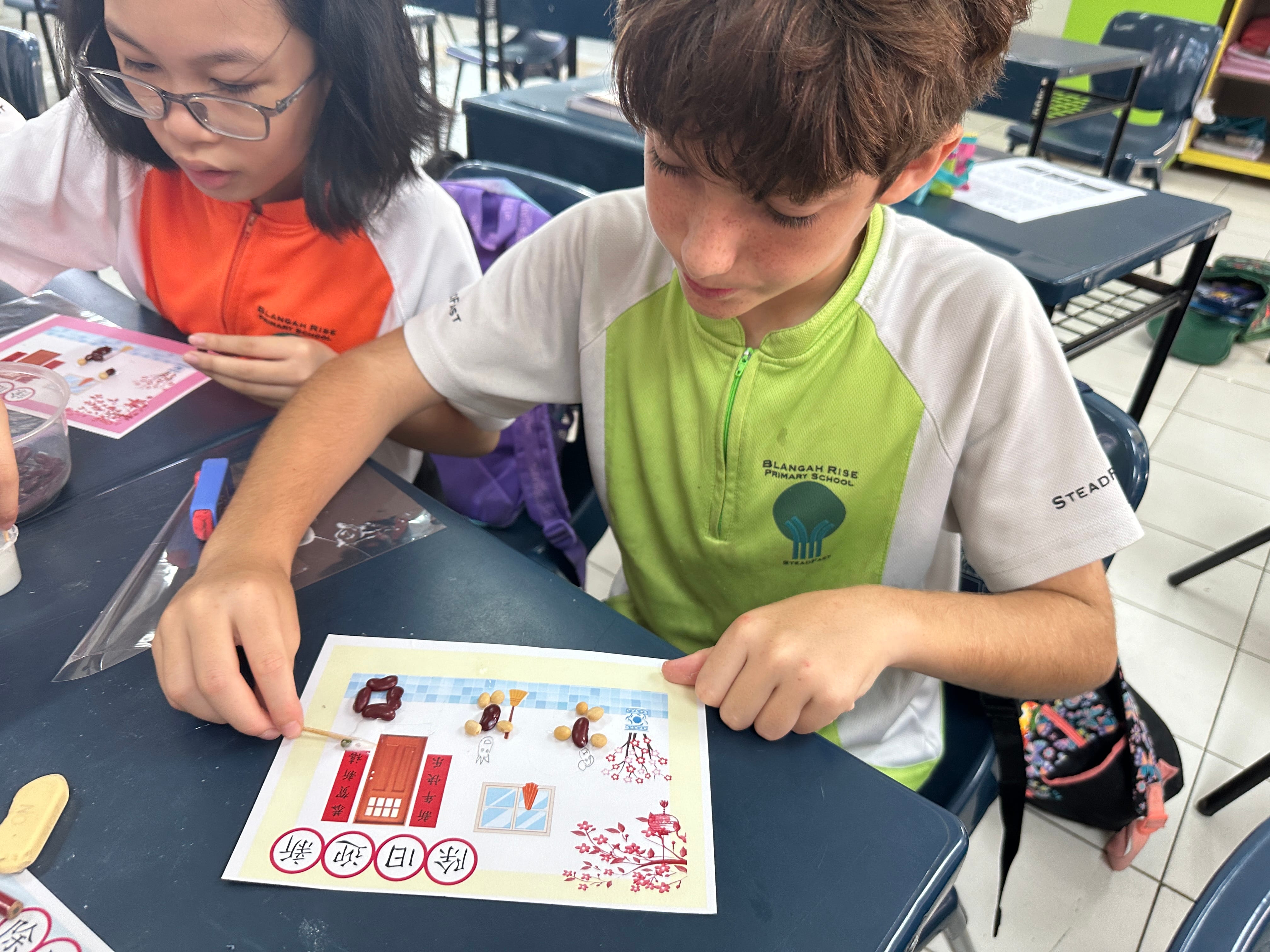 Two primary school students in uniforms work on a craft activity with small objects on a printed sheet with Chinese characters.