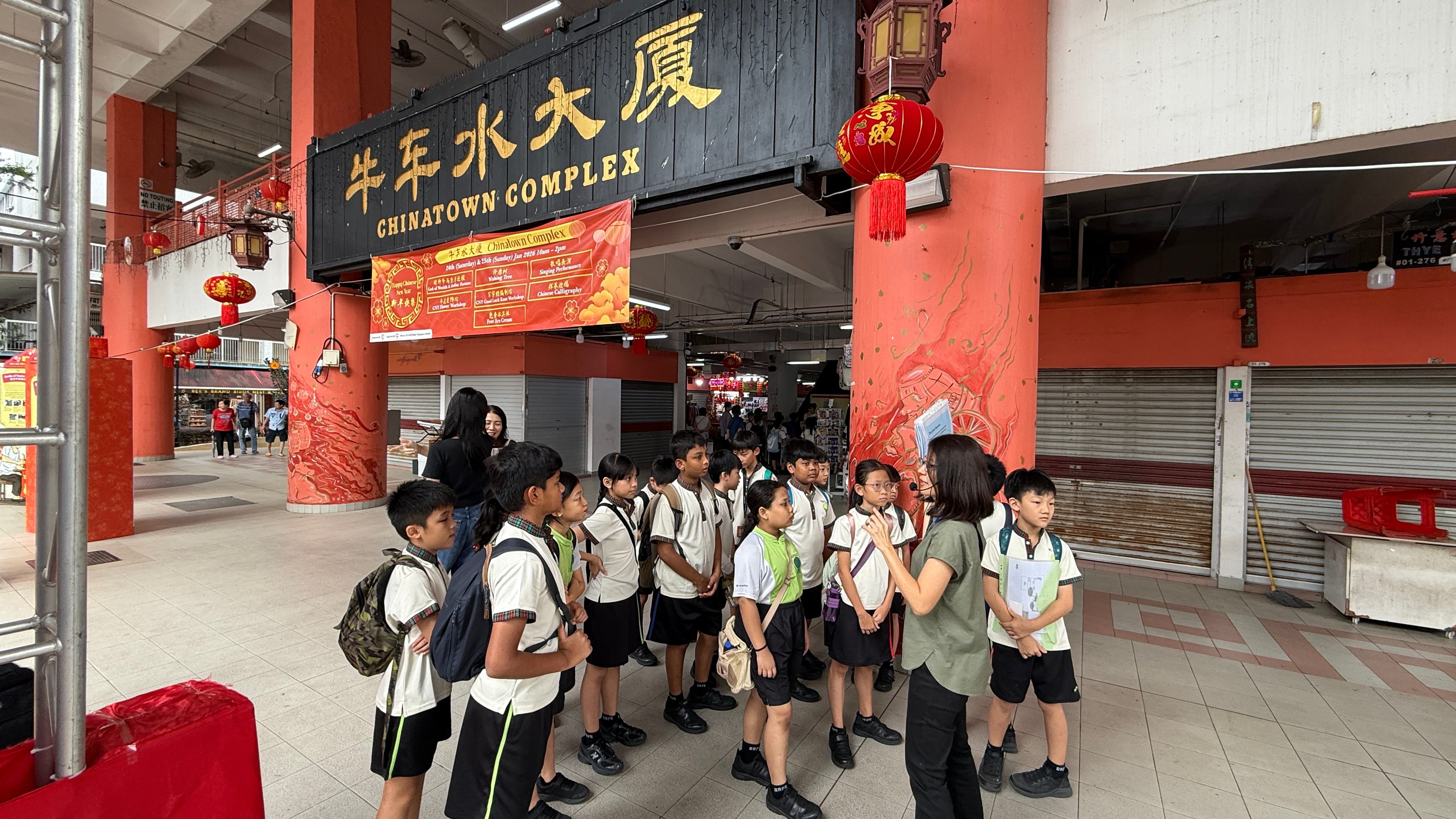 A group of school children in uniforms listen to a guide outside Singapore's Chinatown Complex, decorated with red lanterns.