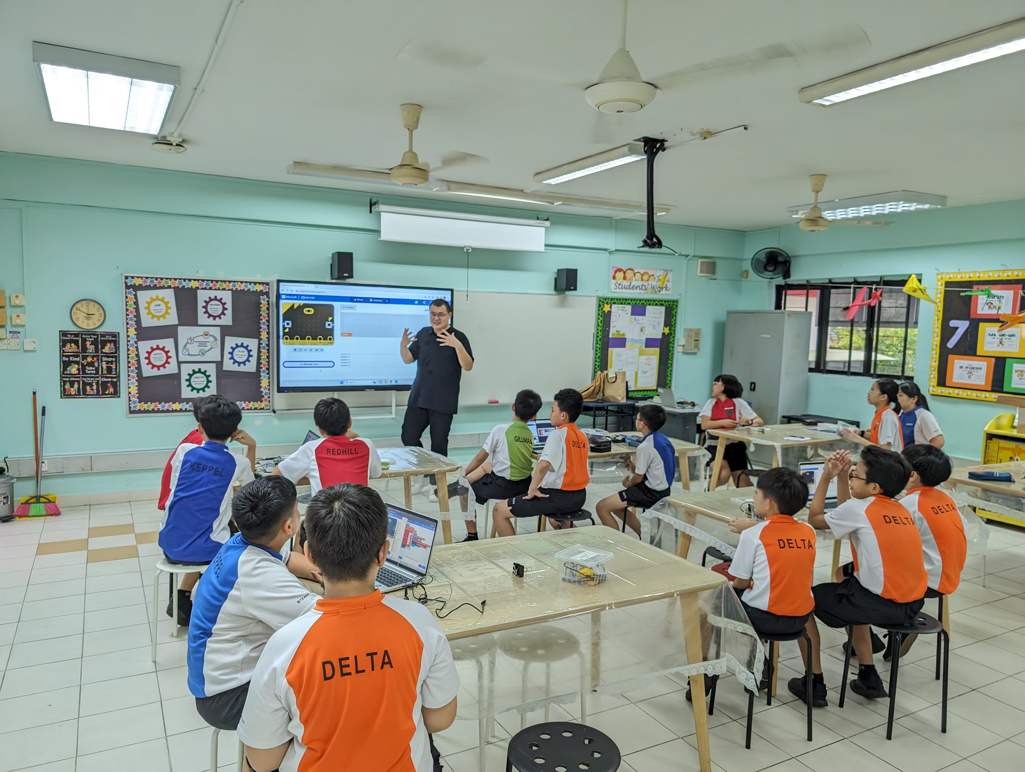 A teacher stands before a screen presenting to young students in orange and white Delta uniforms seated at tables with laptops.