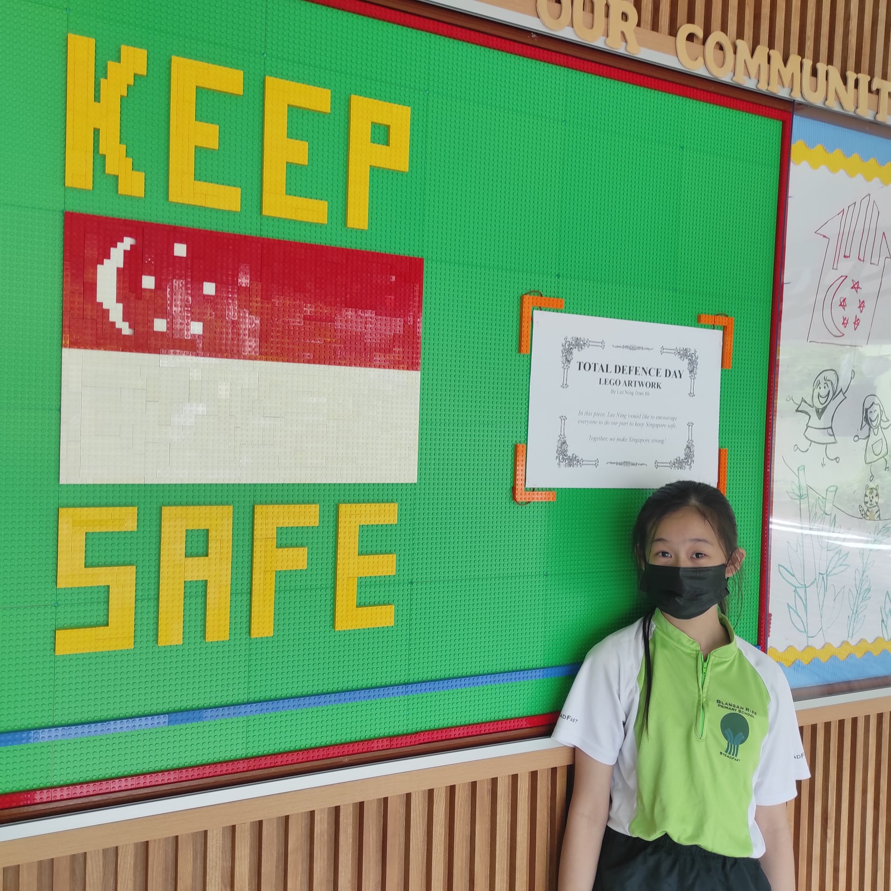 A masked girl in school uniform stands beside a large Lego wall displaying "Keep Safe" and the Singapore flag.