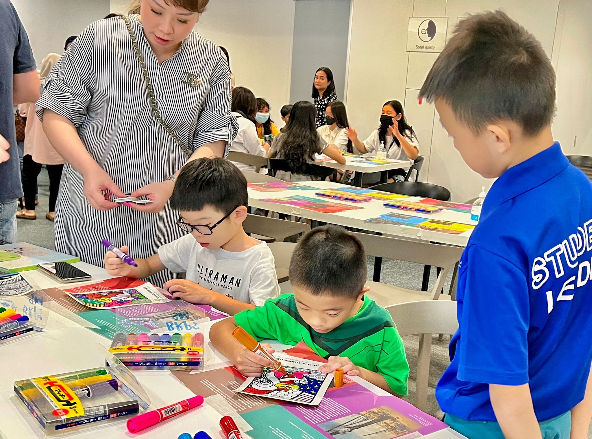 Three children color with markers at a table while an adult woman in a striped dress supervises indoors.