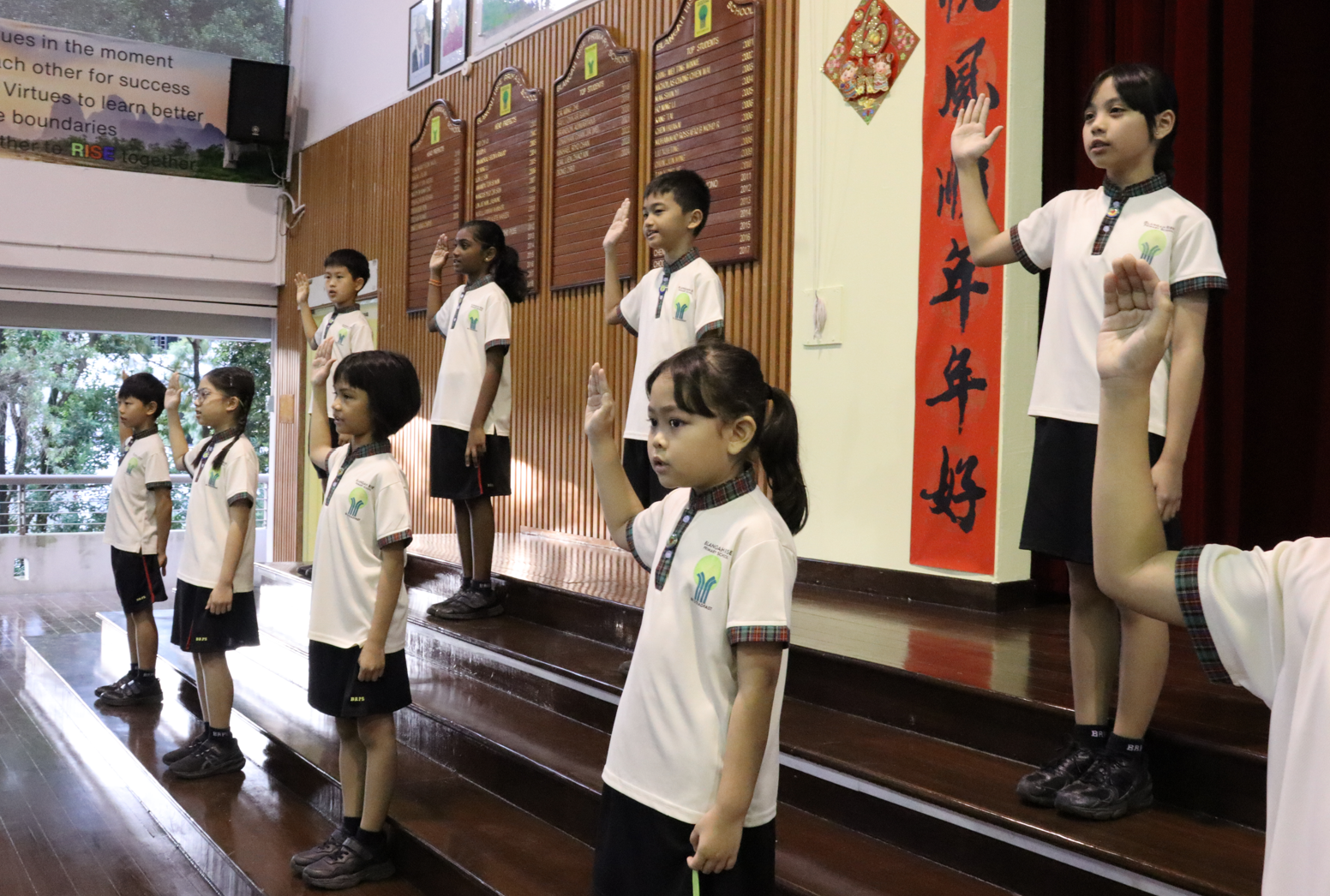 Seven school children in white uniforms raise their right hands on tiered steps beside a red Chinese banner.