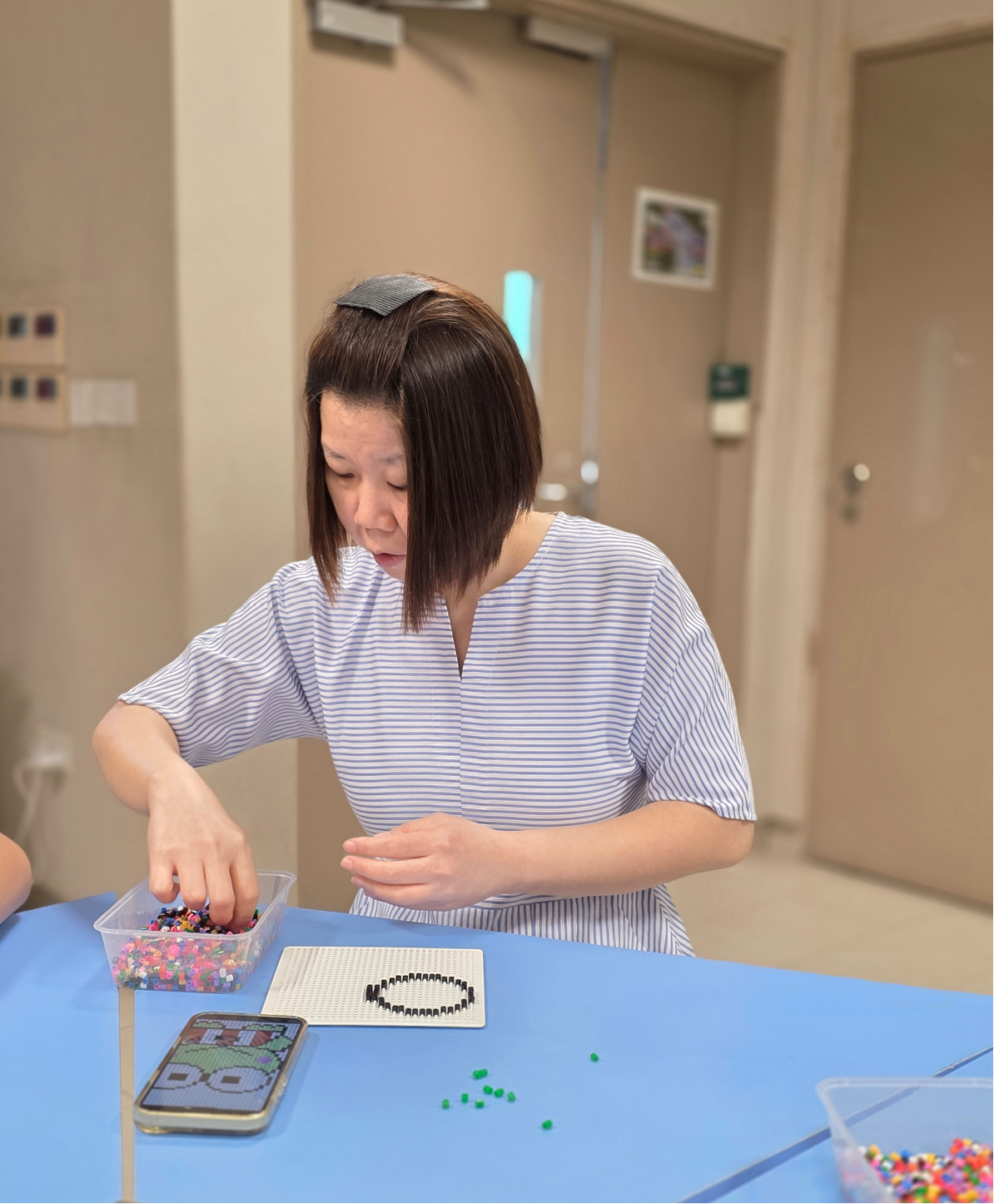 Woman with short dark hair placing colorful beads on a pegboard at a blue table, crafting a bead art design.