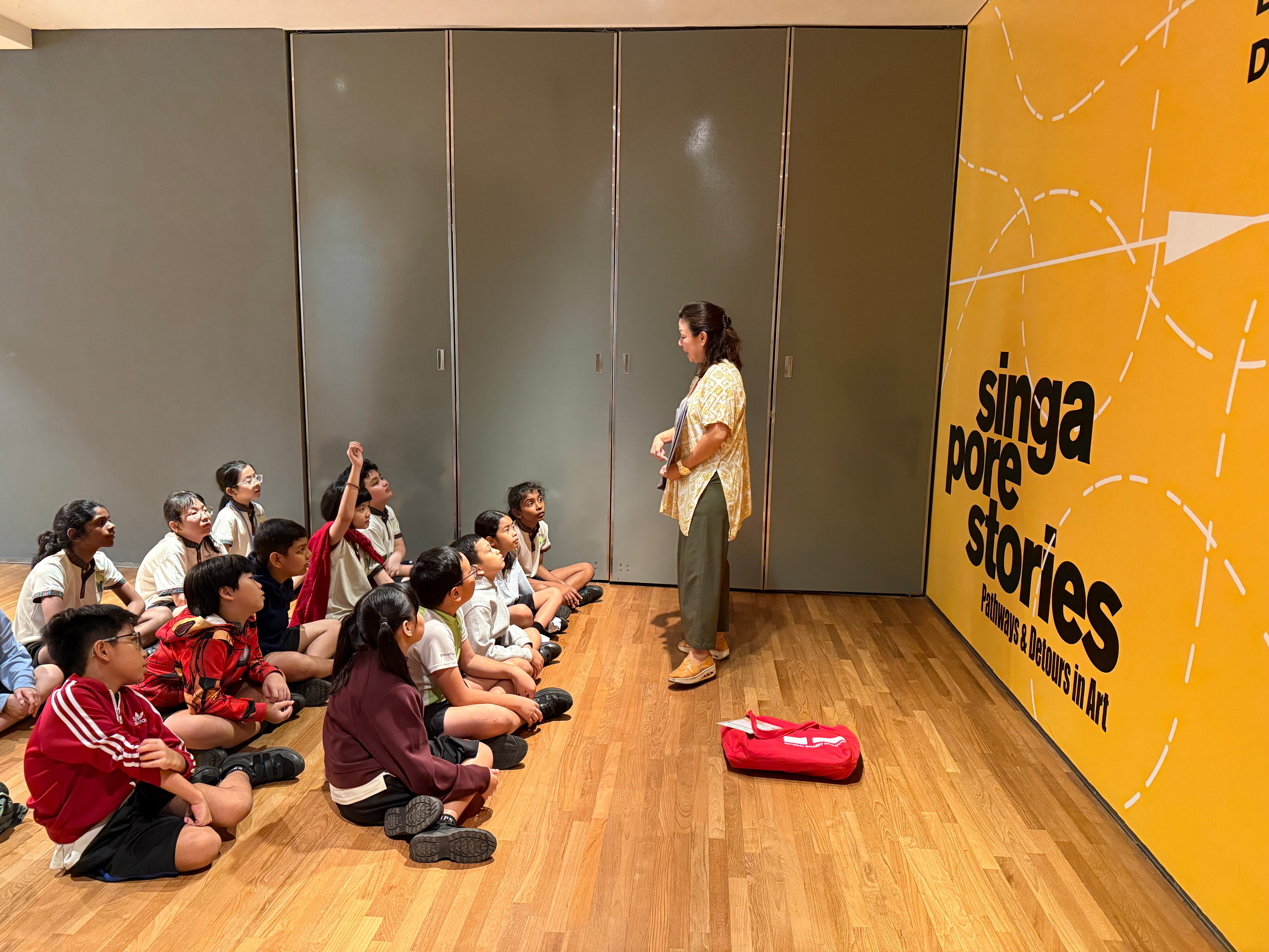 Teacher stands before seated students beside yellow wall reading Singapore Stories; red bag on wooden floor.