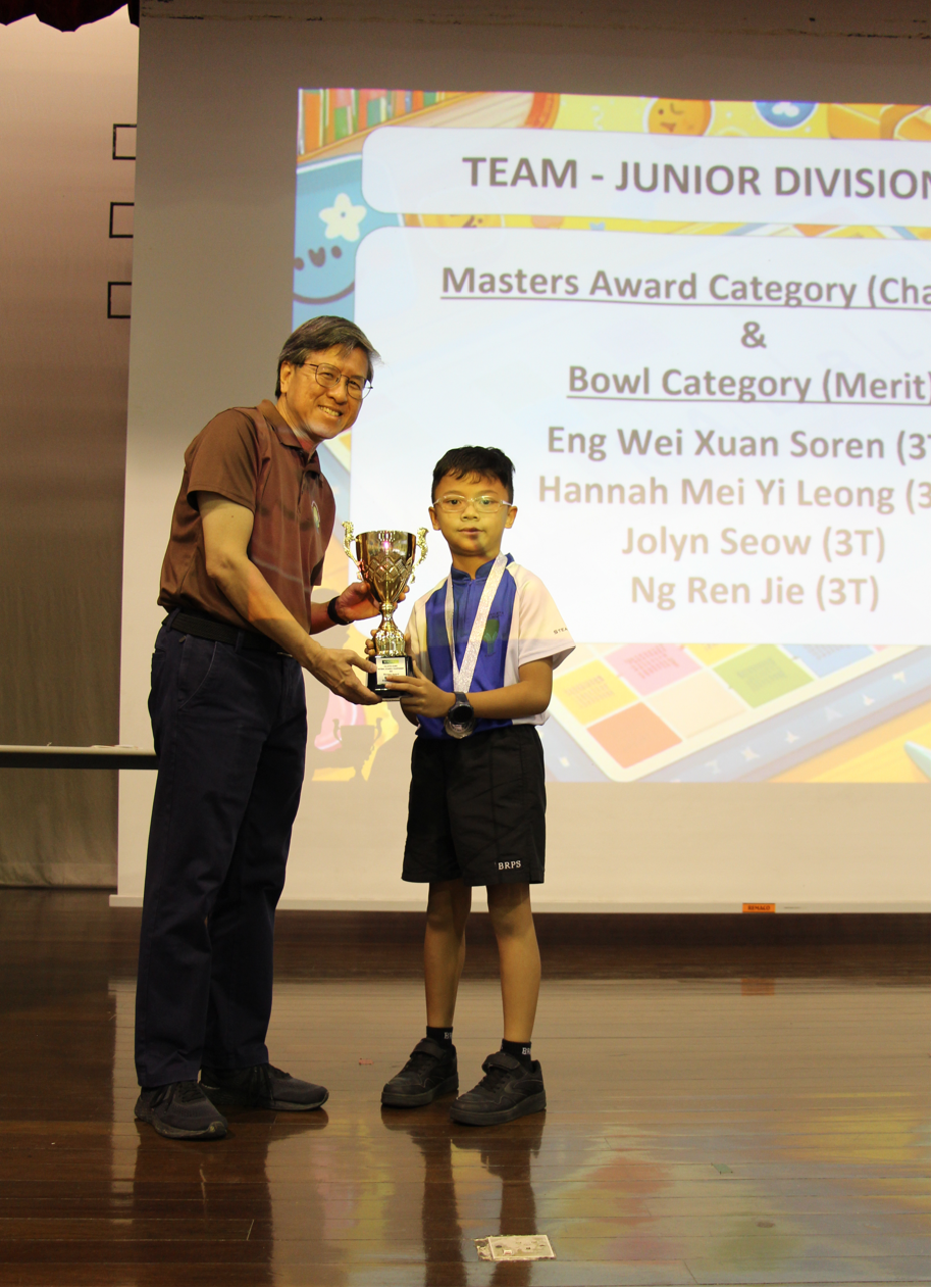 An adult man presents a trophy to a young boy on stage, with a Junior Division Masters Award slideshow behind them.