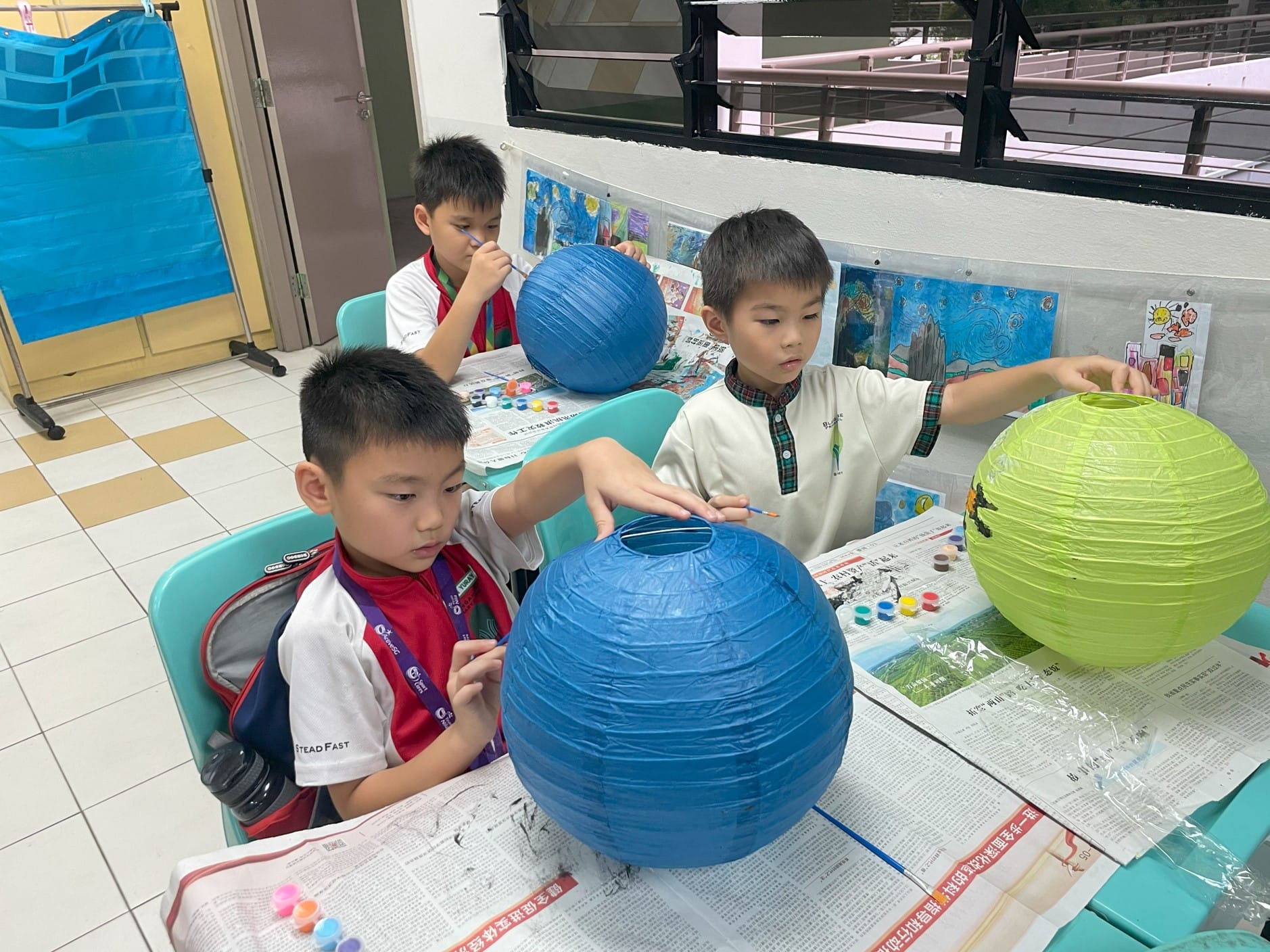 Three boys painting large round paper lanterns in blue and green at a table covered with newspaper.