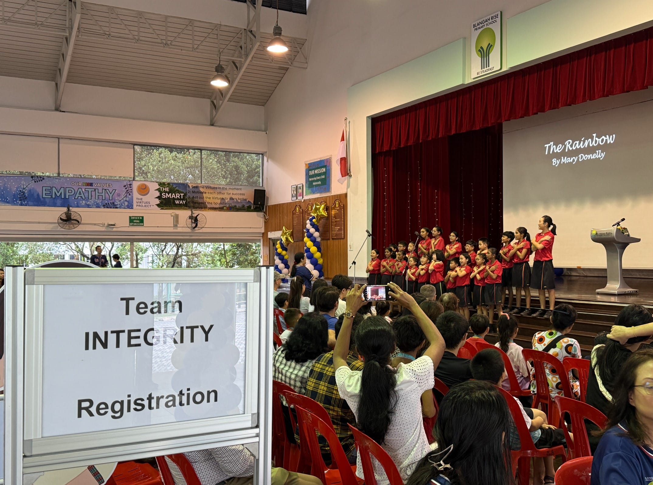 Students in red uniforms perform on a school stage as an audience watches, with a Team Integrity Registration sign in the foreground.