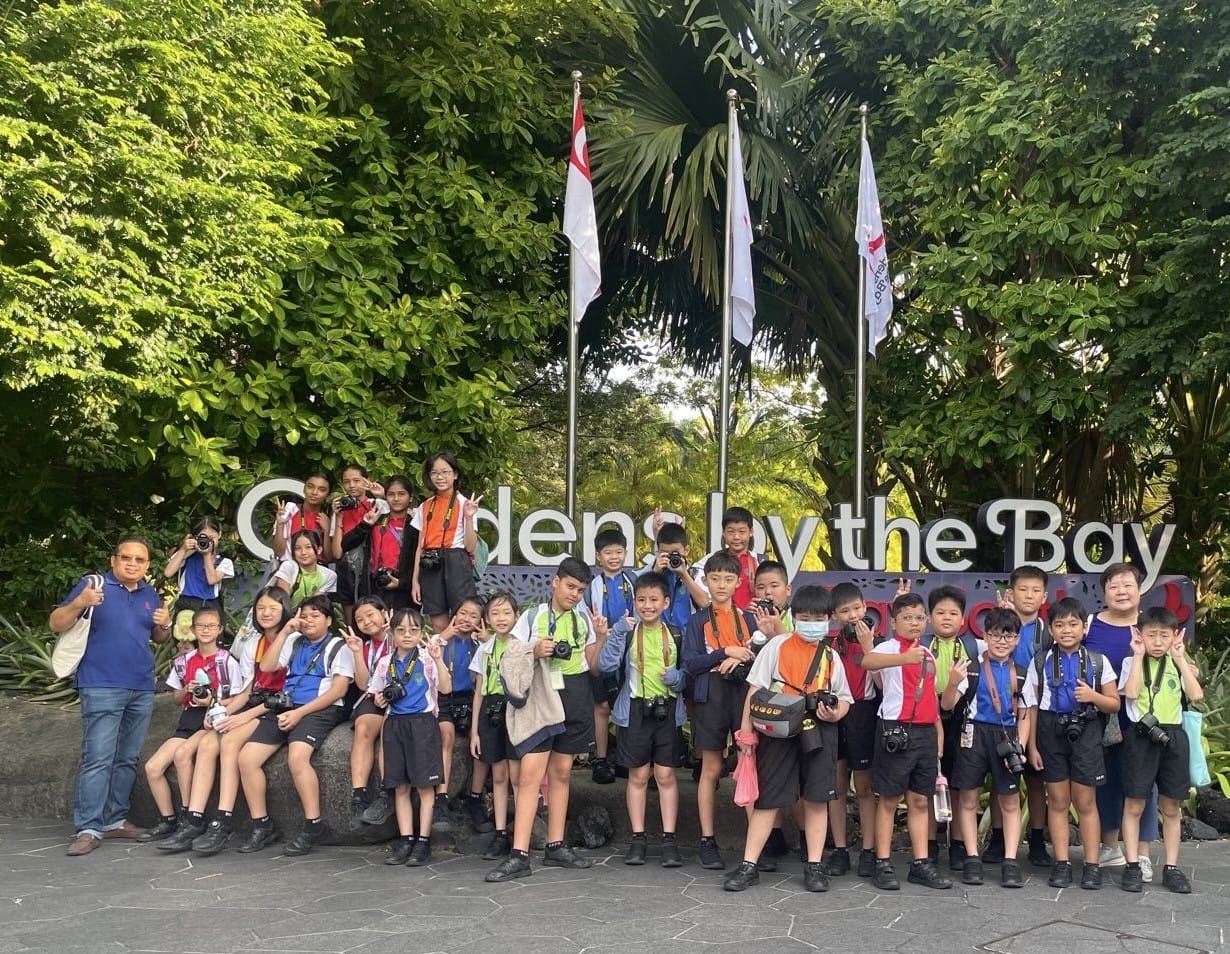 A group of school children with cameras pose in front of the Gardens by the Bay sign in Singapore.